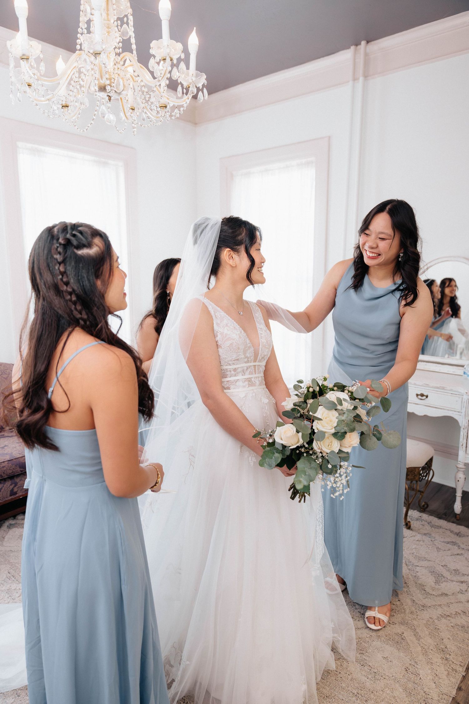 Bride in white gown holds bouquet surrounded by bridesmaids in blue dresses in elegant white room with chandelier.