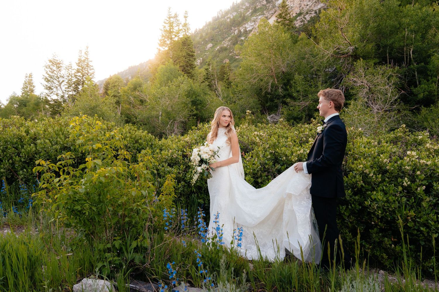 Bride and groom stand among wildflowers with mountain backdrop, groom holding wedding dress train at golden hour.