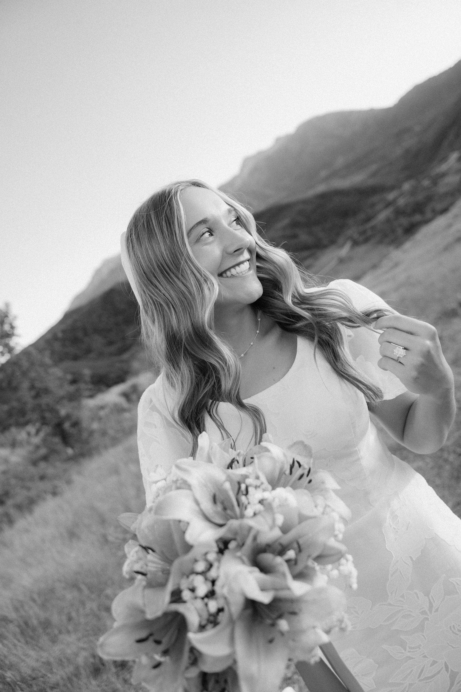Smiling bride holds floral bouquet with mountain backdrop in black and white photo.