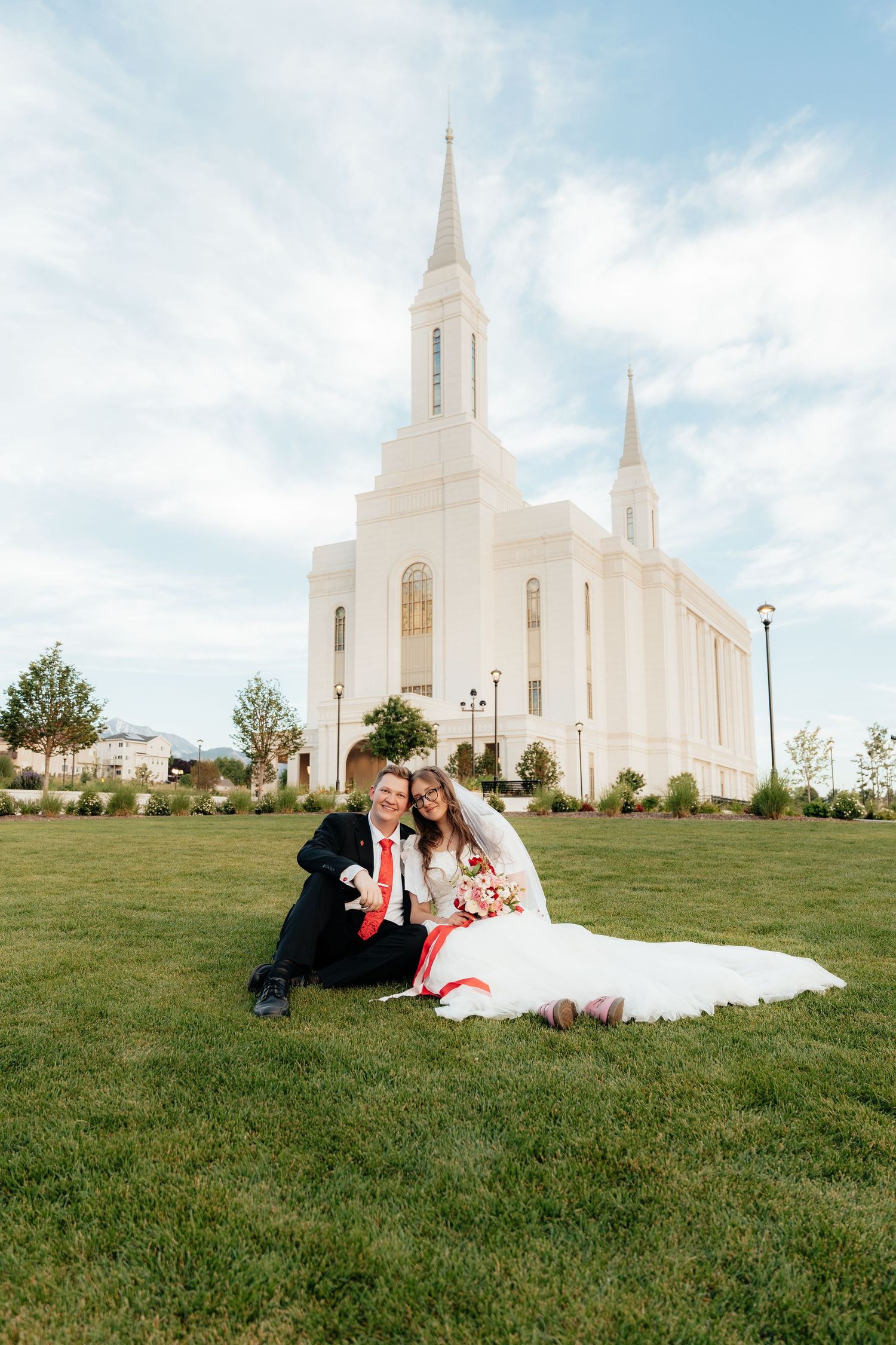 Bride and groom sitting on green grass in front of a white LDS temple with spires on their wedding day.
