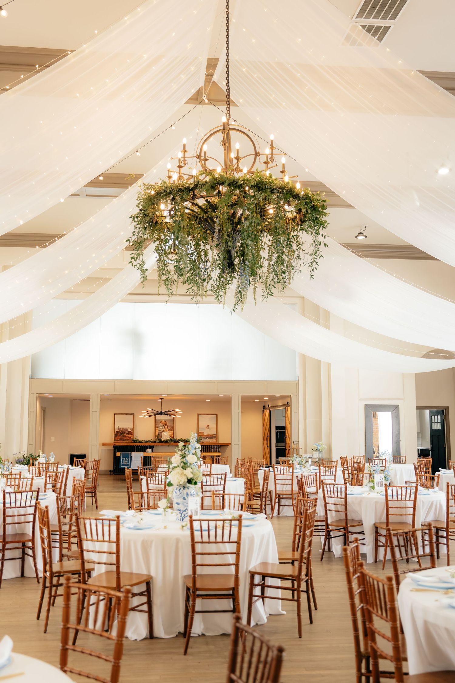 Elegant wedding reception hall with white draping, chiavari chairs, round tables, and a floral chandelier centerpiece.