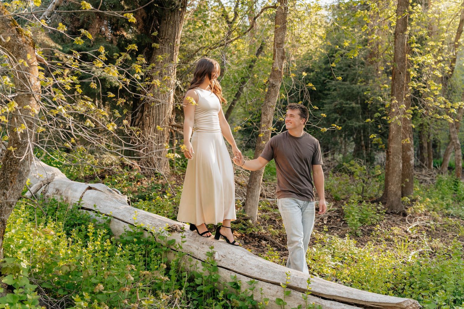 Couple holding hands walking on a fallen log in a sunlit forest wearing casual summer attire.