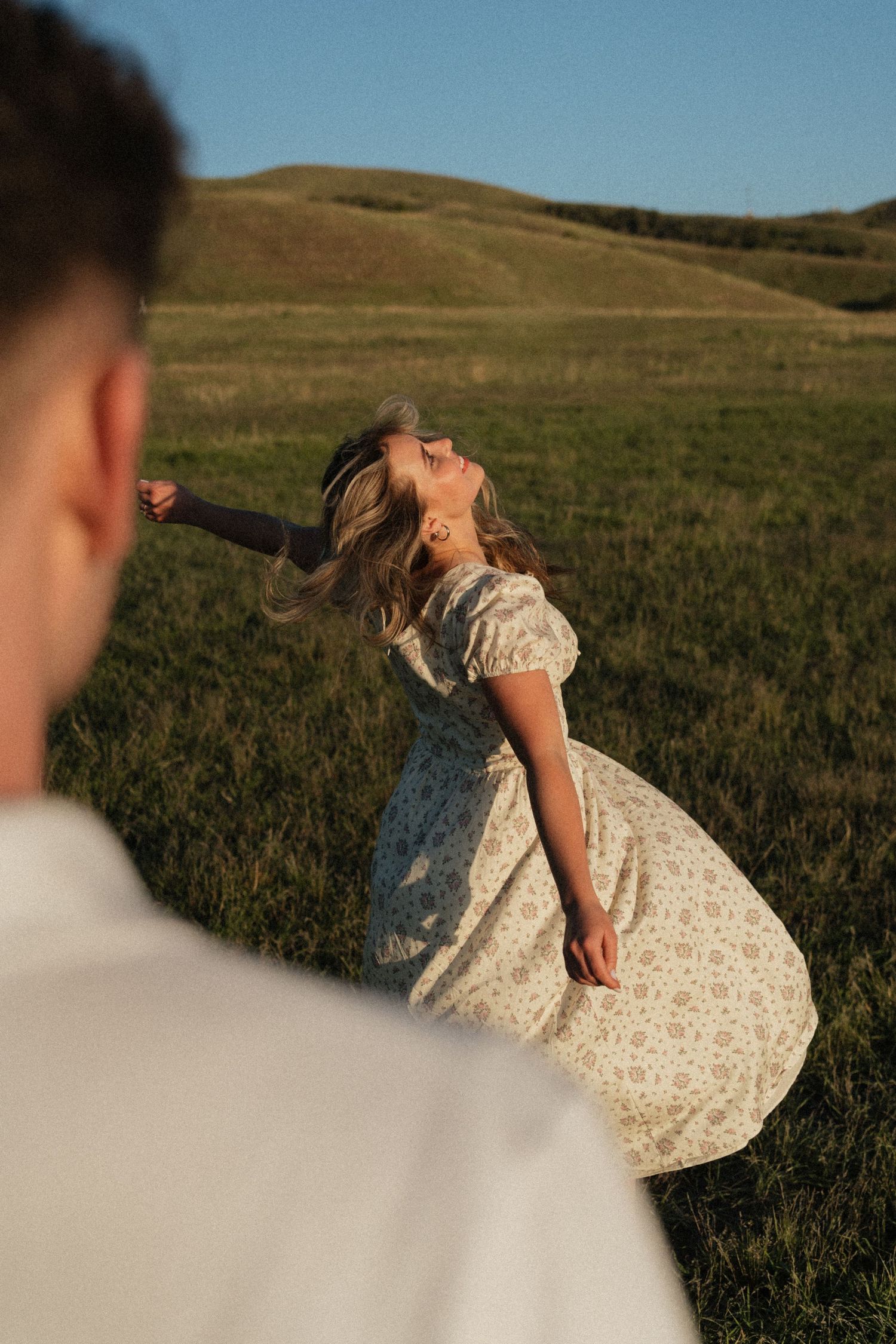 A woman in a floral midi dress twirls joyfully in a green meadow with rolling hills, as a man watches her.