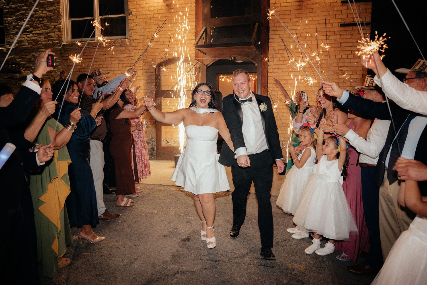 Bride and groom exit wedding venue through sparkler tunnel surrounded by cheering guests at night.