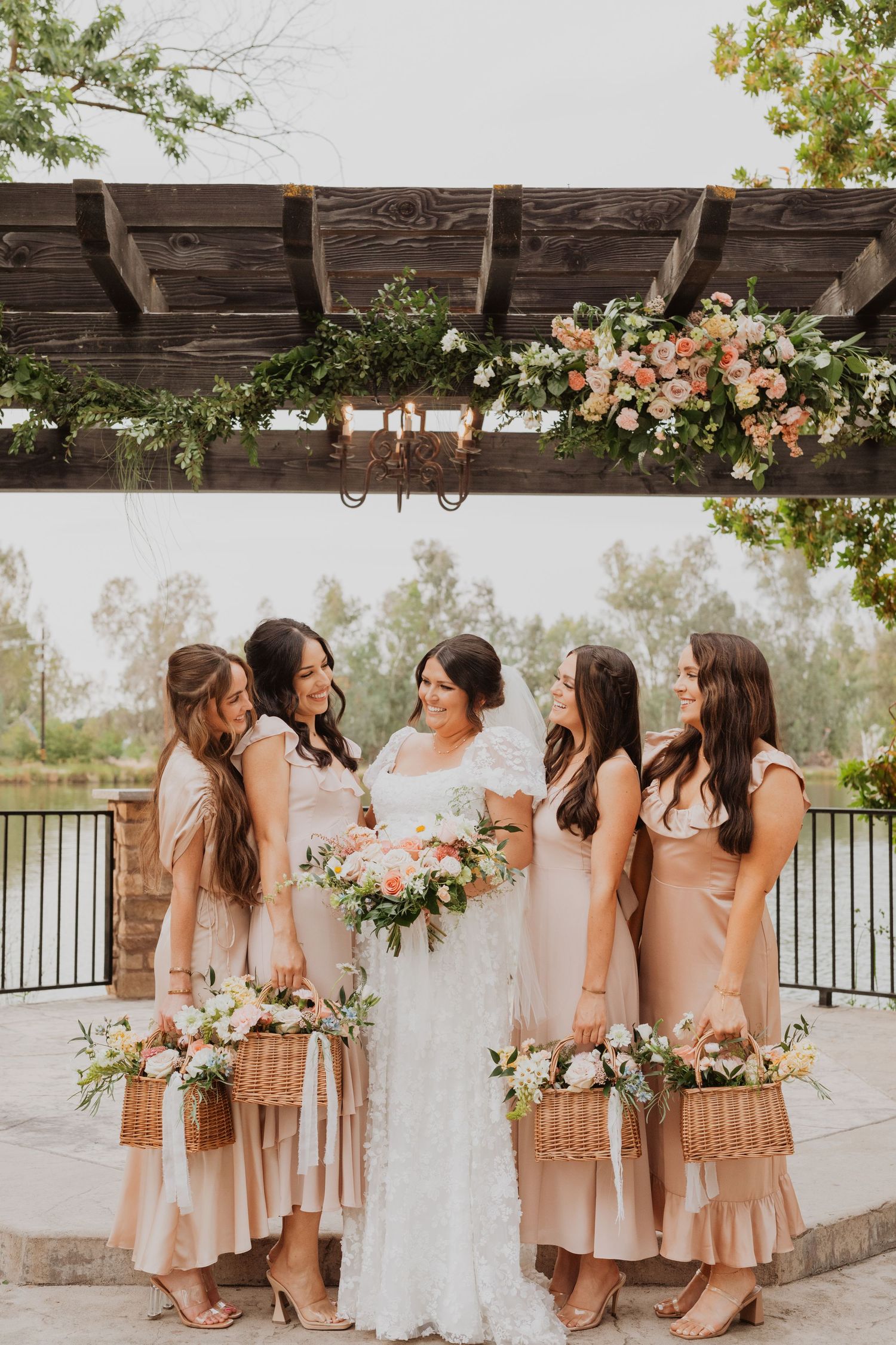 Wedding party in neutral beige dresses stands together on a stone patio with floral decorations overhead.
