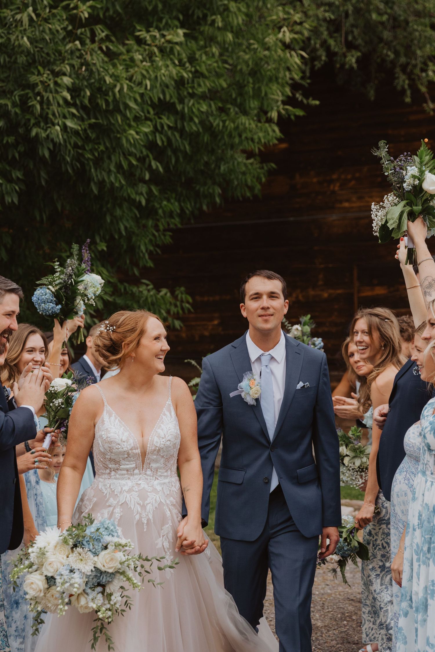 Wedding party celebrates outdoors wearing navy suits and light blue dresses against a rustic backdrop.