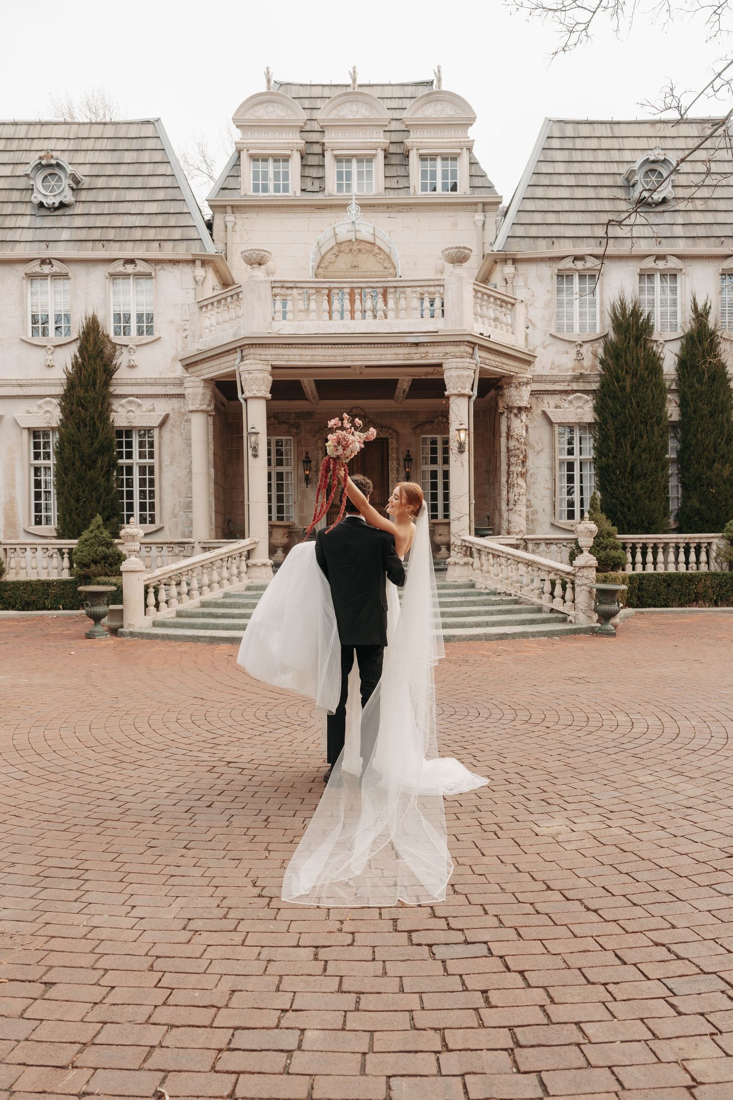 A couple shares a romantic moment outside an elegant French-style mansion with cypress trees and brick courtyard.