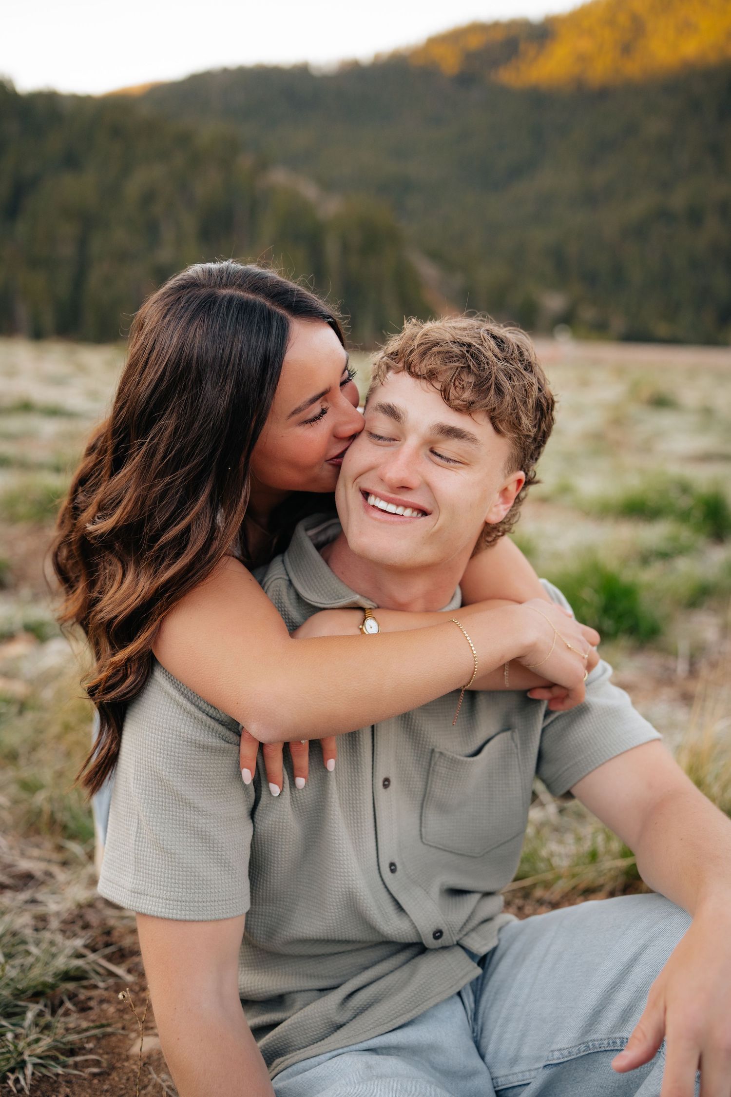 A young couple shares a romantic moment outdoors, with the woman kissing the smiling man on the cheek in a scenic meadow.