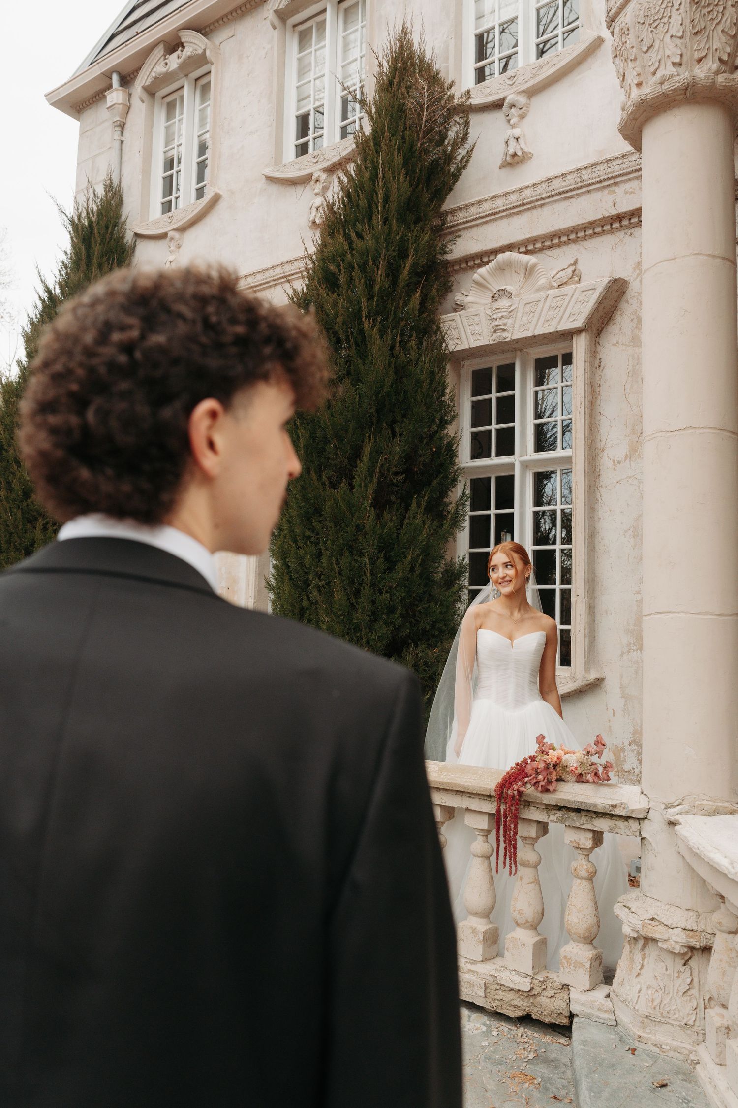 A romantic wedding moment captured at an elegant mansion with tall cypress trees and ornate stone balustrades.