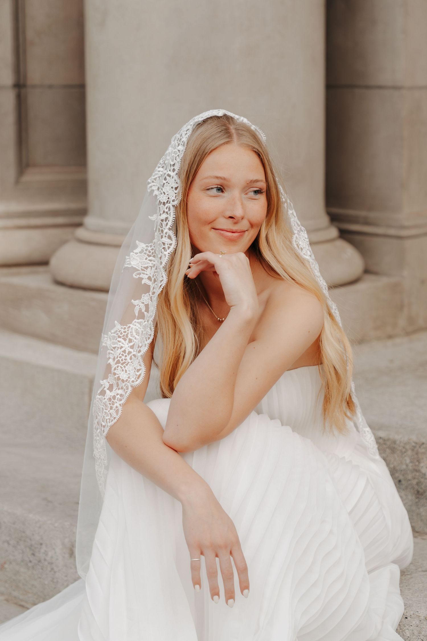 A bride in a white lace veil and wedding dress poses elegantly against stone columns in a romantic bridal portrait series.