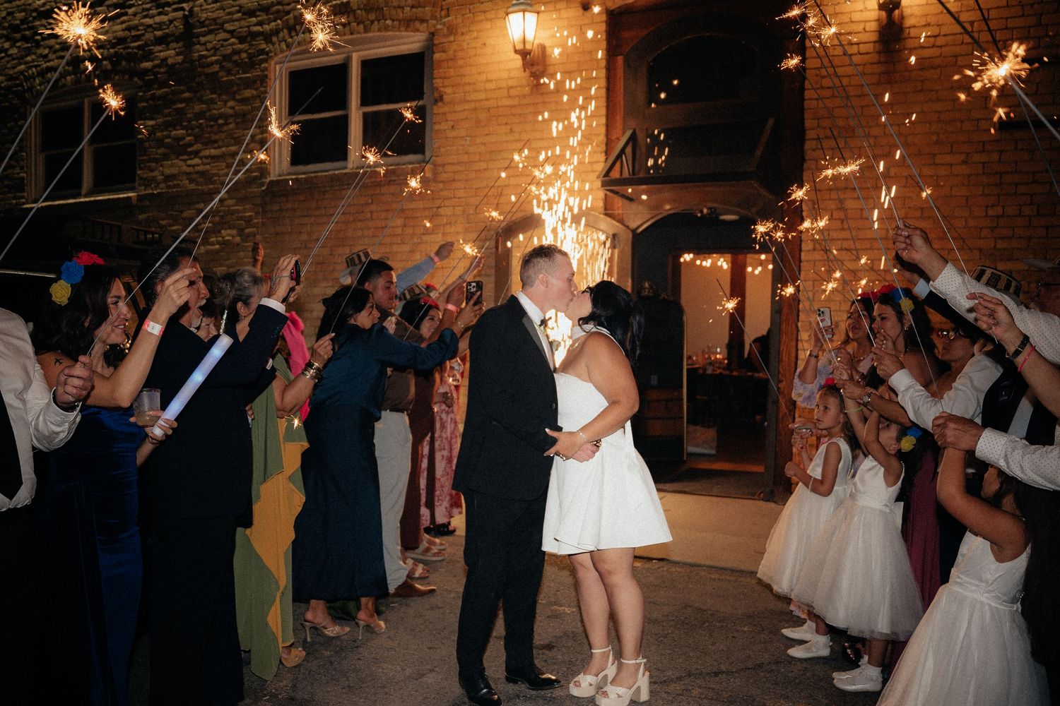 Bride and groom share a kiss during a sparkler send-off surrounded by cheering wedding guests at night.