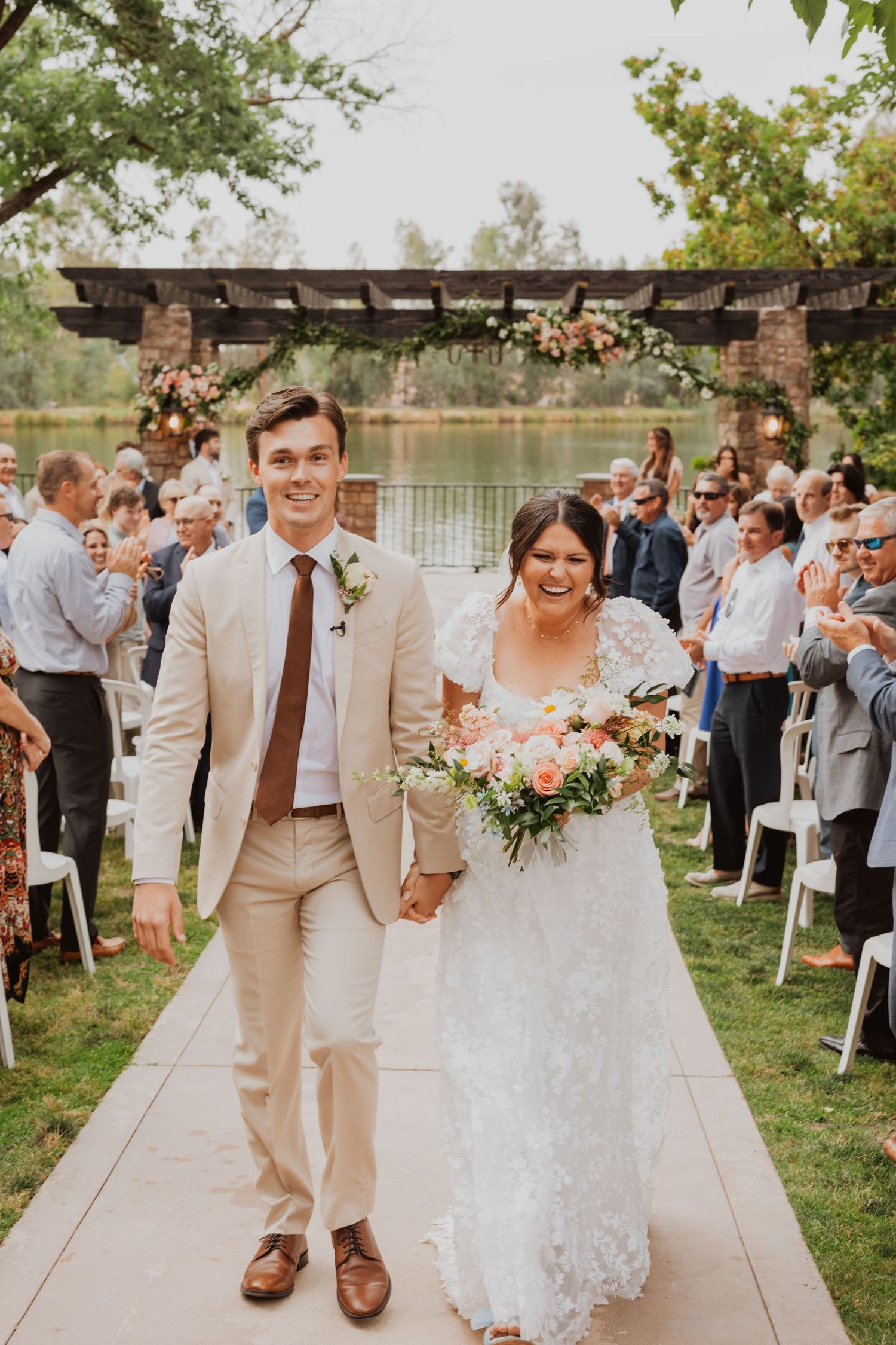 Wedding party walks along waterfront path with guests gathered in background.