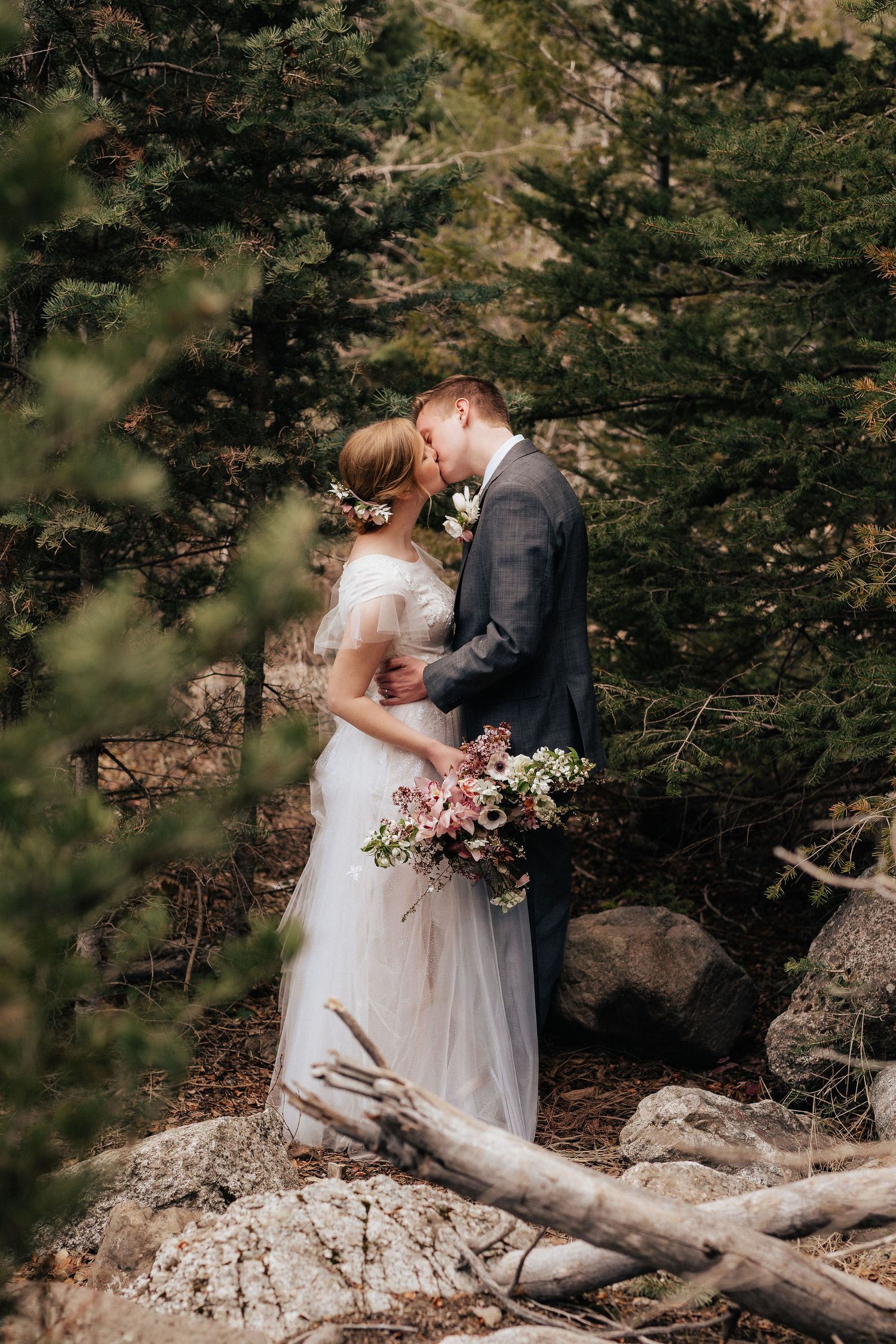 Intimate wedding moment captured in a forest setting with evergreen trees and natural rocks.