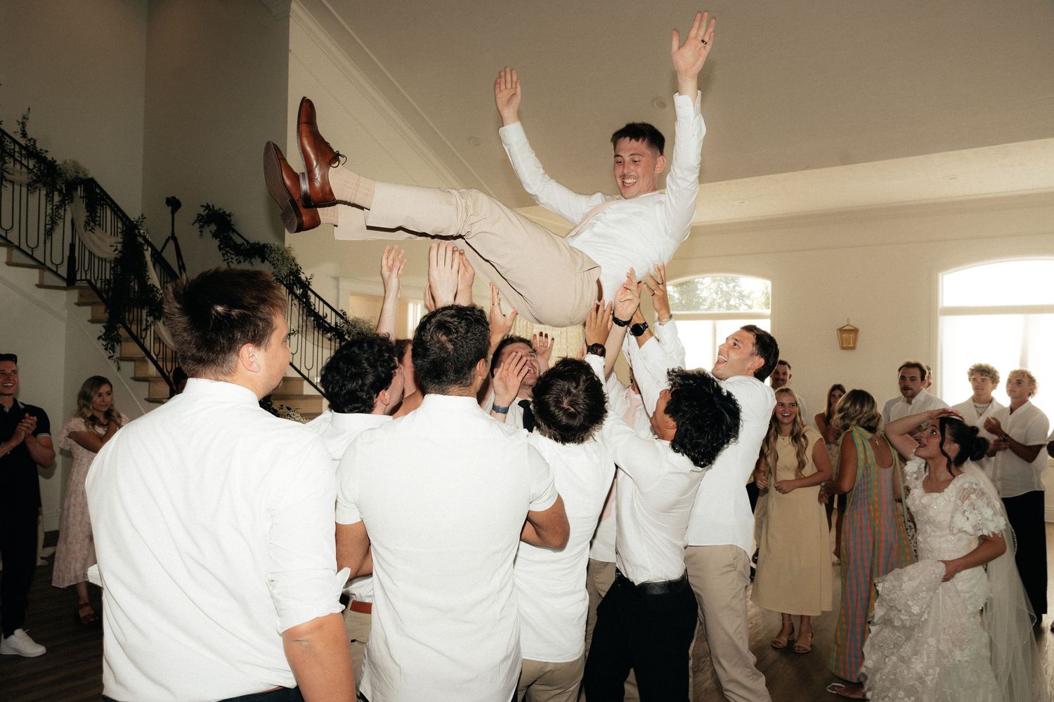 Groom in white suit tossed in air by groomsmen during lively wedding reception celebration indoors.