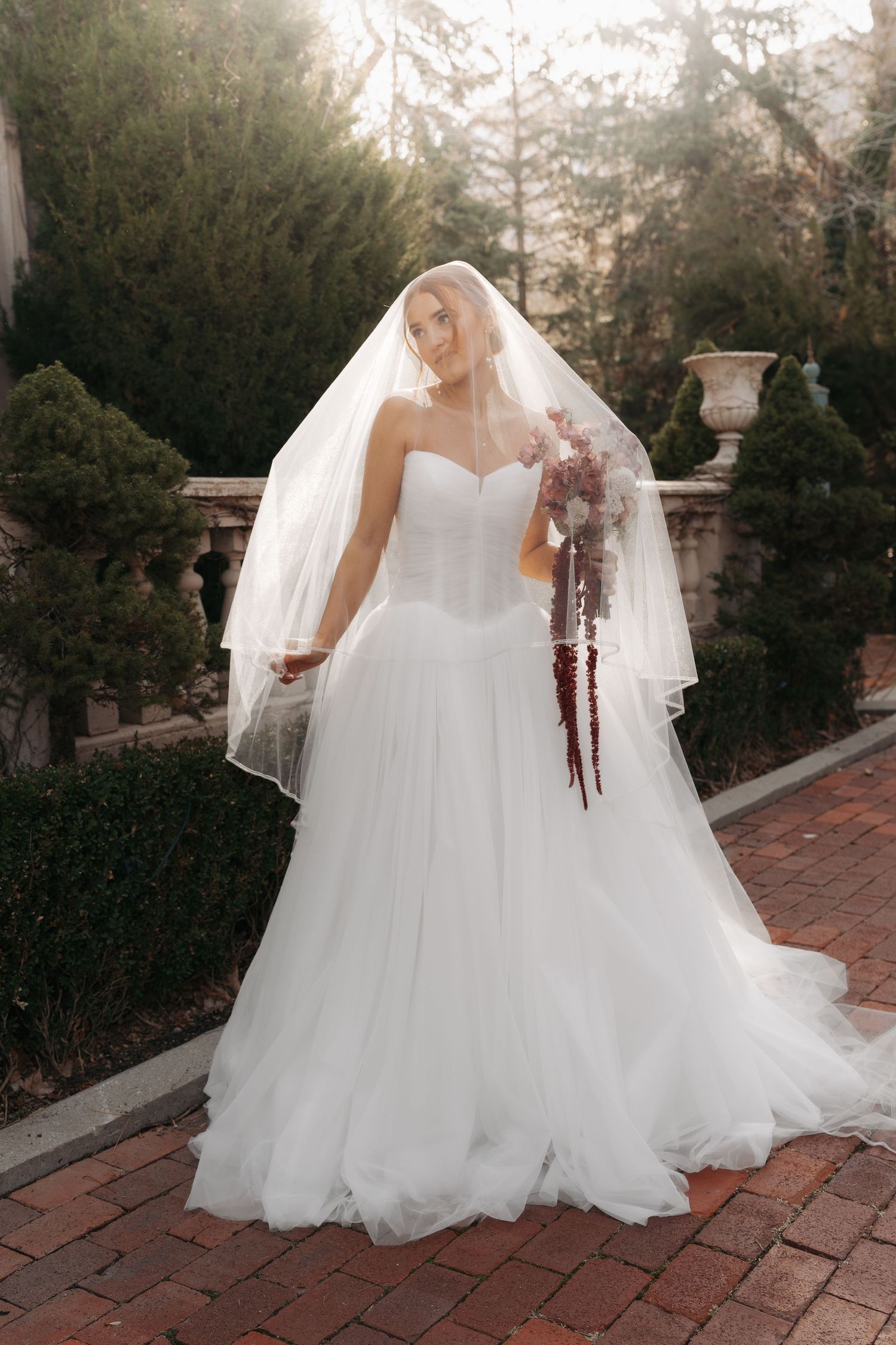 A bride in a flowing white strapless gown and veil poses in a garden setting with a cascading burgundy bouquet.