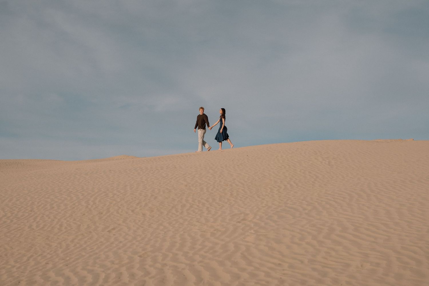 Two distant figures walk along the crest of a rippling sand dune under an overcast sky.