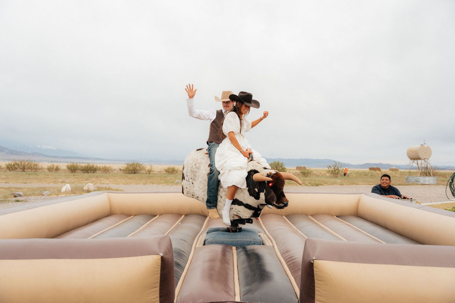 Bride and groom riding a mechanical bull at an outdoor western-themed wedding reception under cloudy skies.