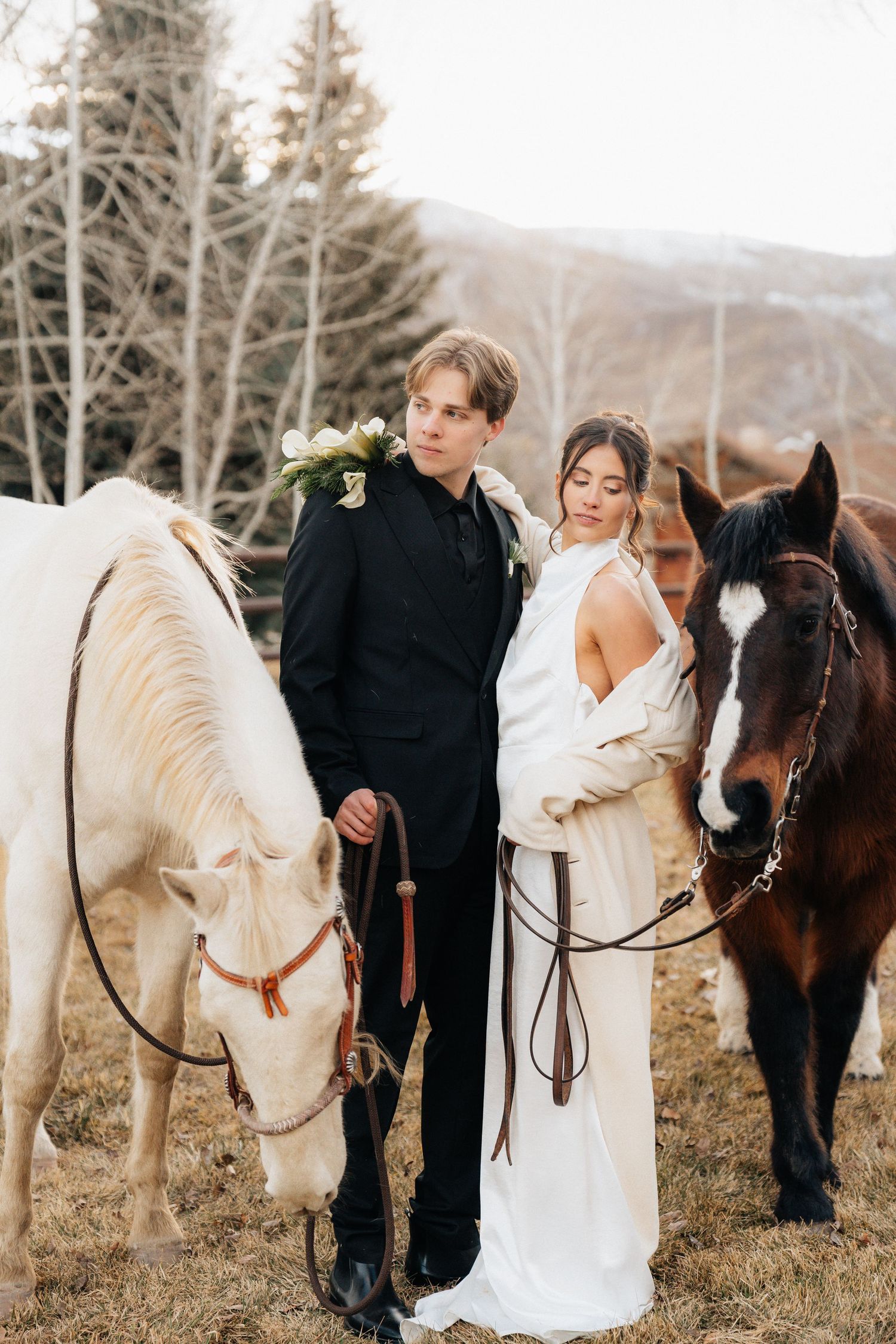 Wedding couple stands between two horses in a field with bare trees and mountains in the background in Utah.