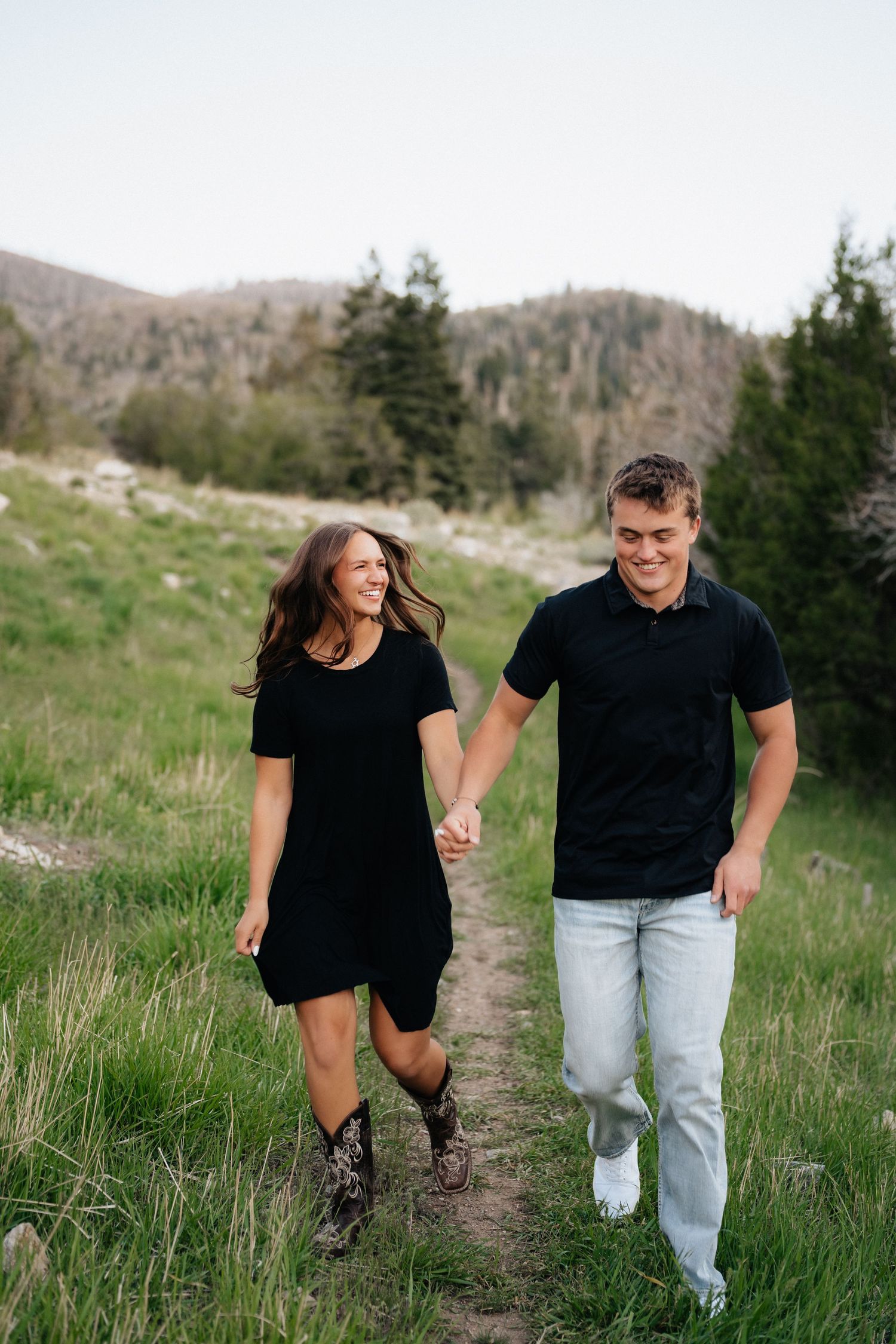 A happy couple holds hands walking on a mountain trail, both wearing black tops, surrounded by green meadows.