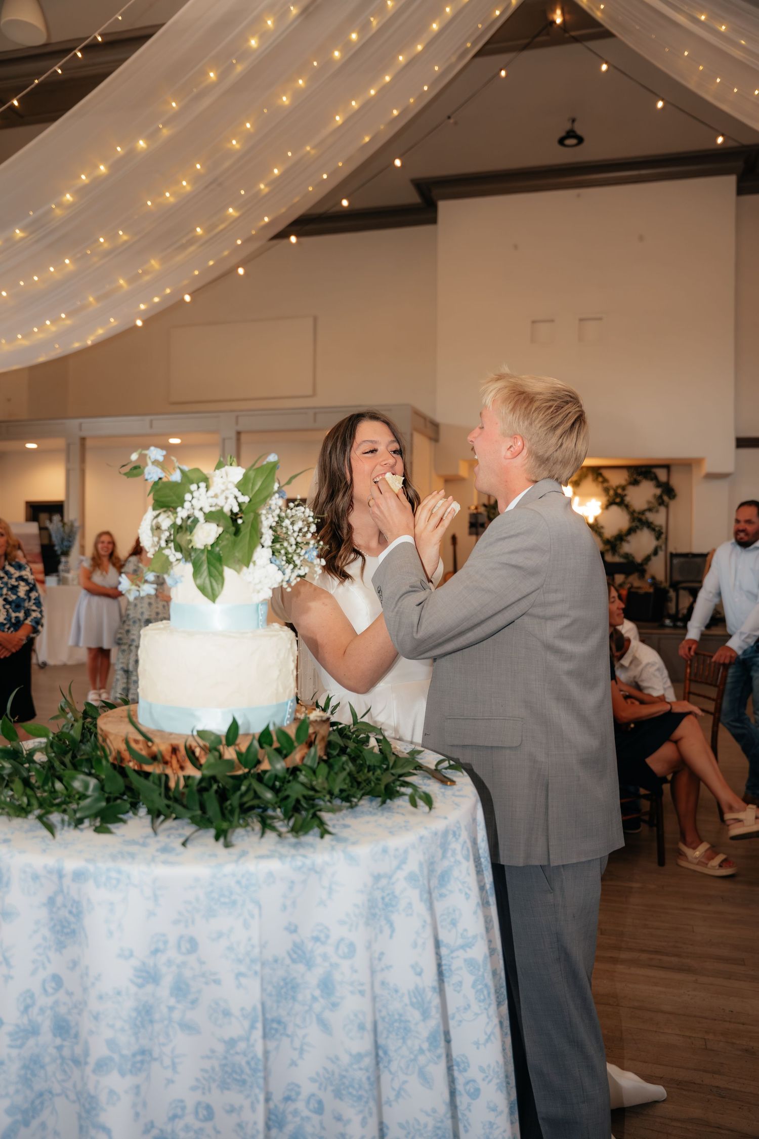 Bride and groom feeding each other wedding cake at reception with fairy lights and floral decorations.
