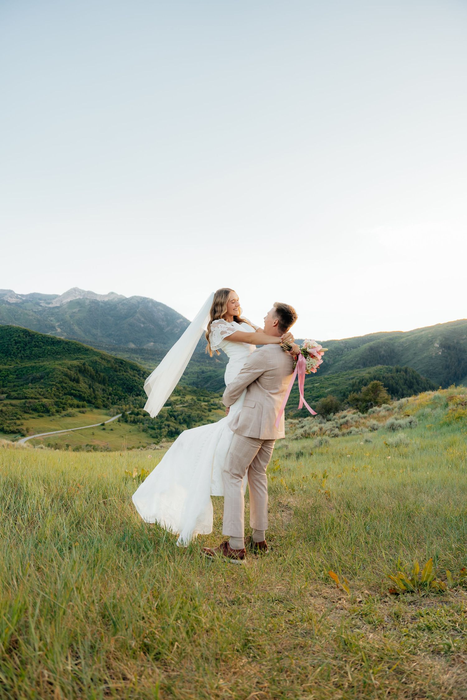 Groom lifting bride in white dress and veil on green mountain hillside during outdoor wedding celebration.