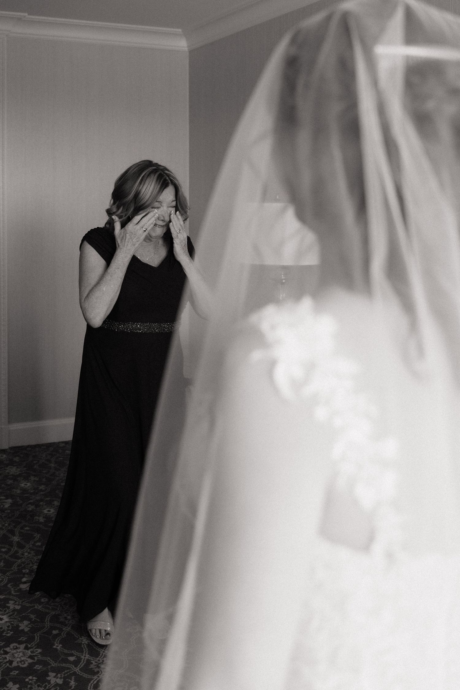 Black and white photo of emotional woman in black dress wiping tears seeing bride in wedding gown and veil.