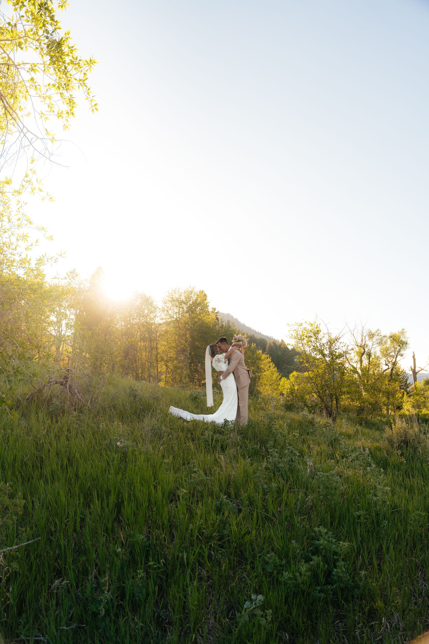 Bride and groom embrace in a sunlit mountain meadow with lush green grass and trees at golden hour.