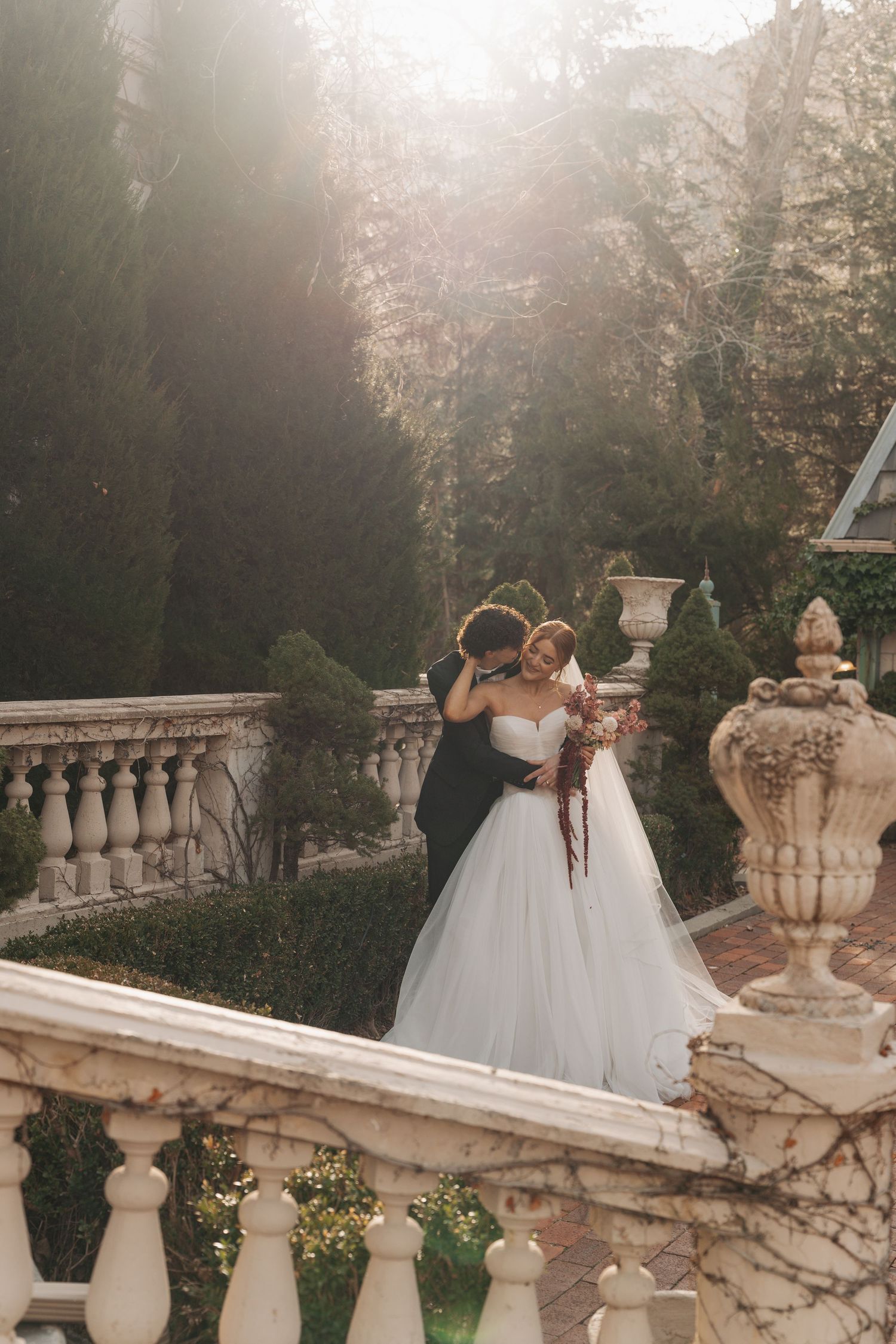 Wedding couple embraces on an ornate stone terrace surrounded by winter gardens at sunset.