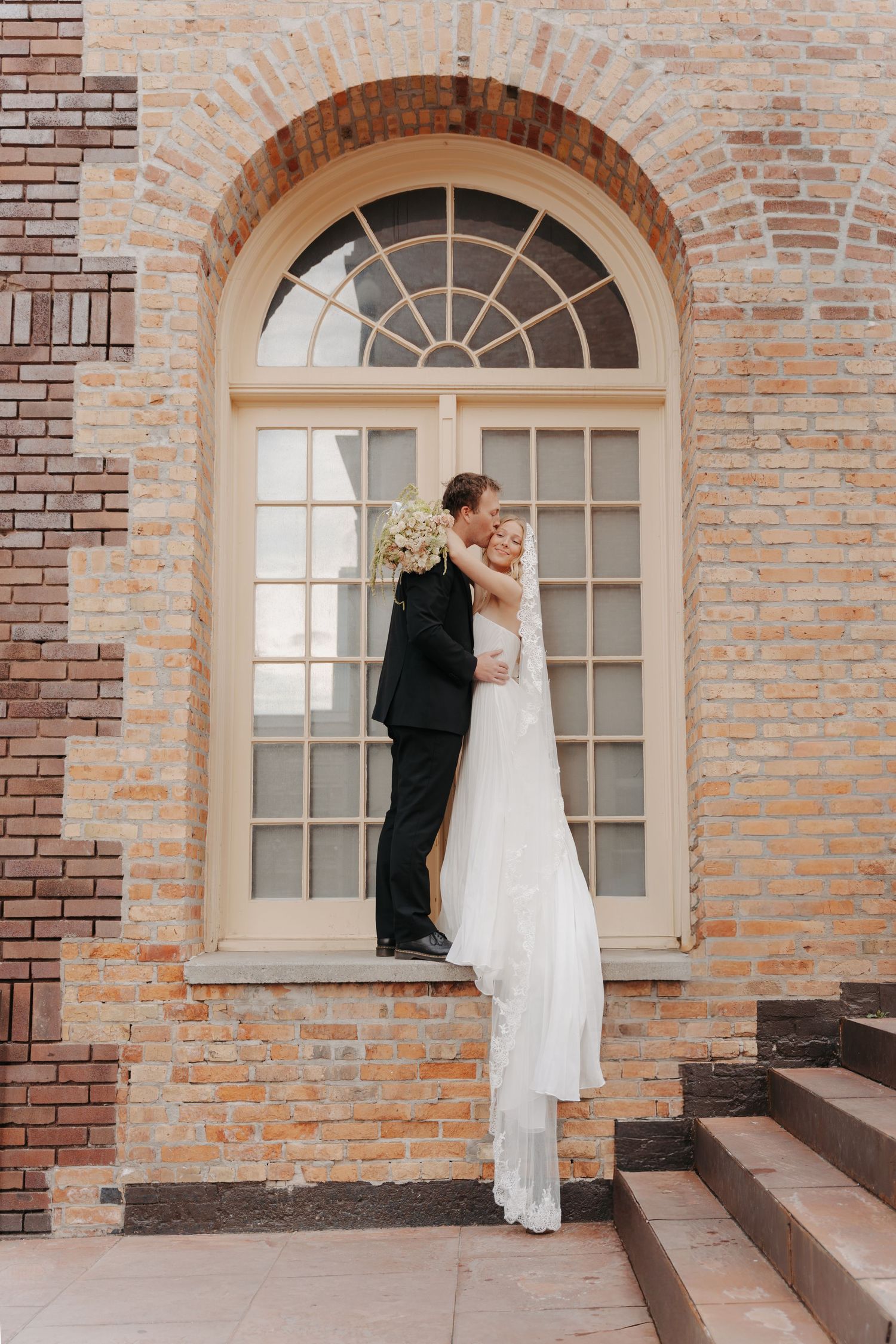 A romantic moment between newlyweds on brick steps with arched windows and white dress flowing in the breeze.