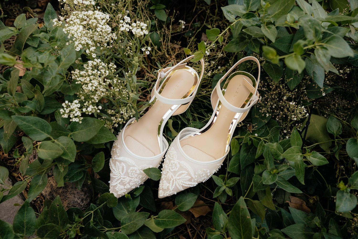 Elegant white lace bridal flats with ankle straps resting on green foliage and delicate white flowers.