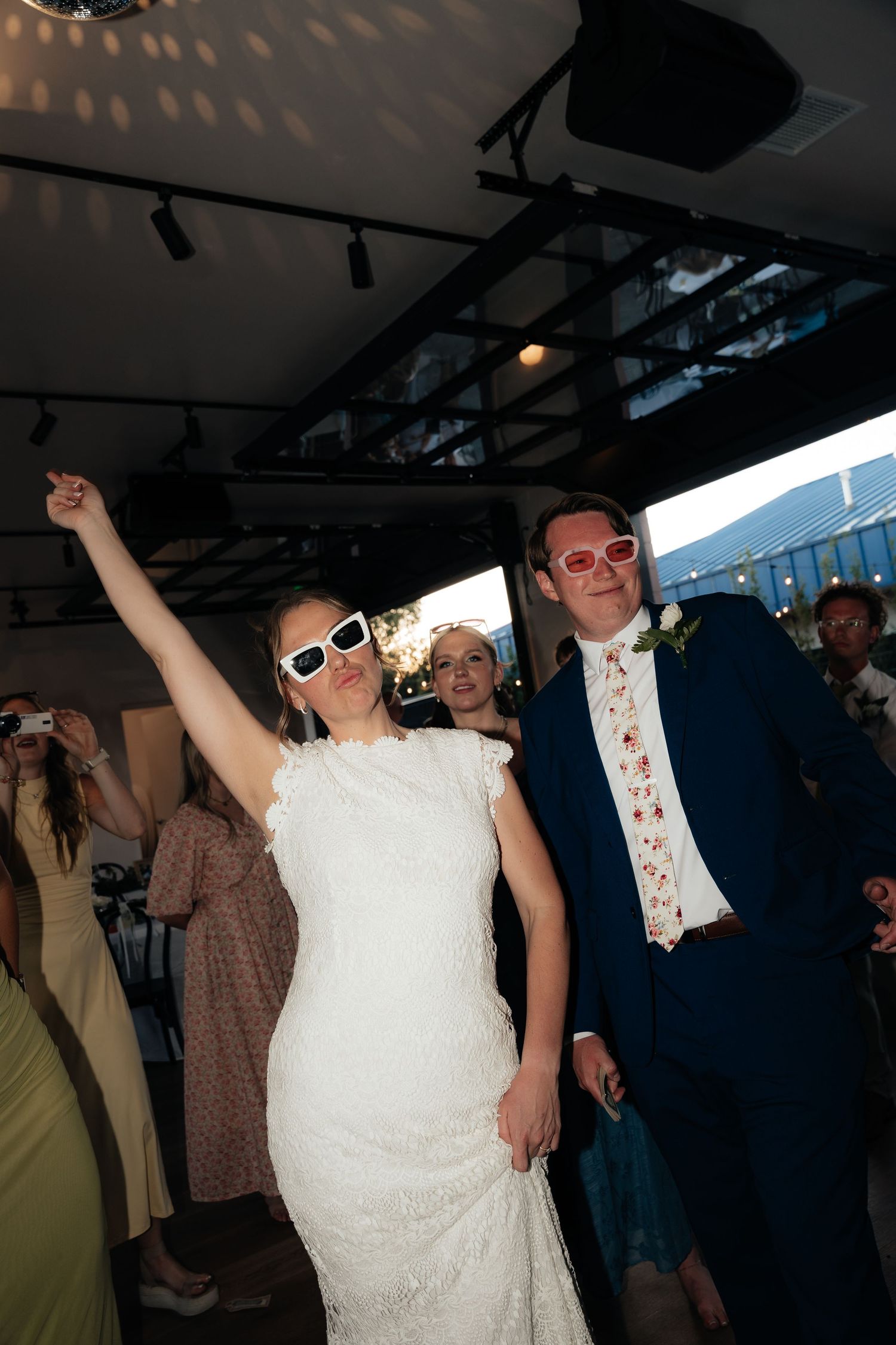 Bride in white lace dress and groom in navy suit wearing sunglasses, dancing joyfully at their wedding reception.