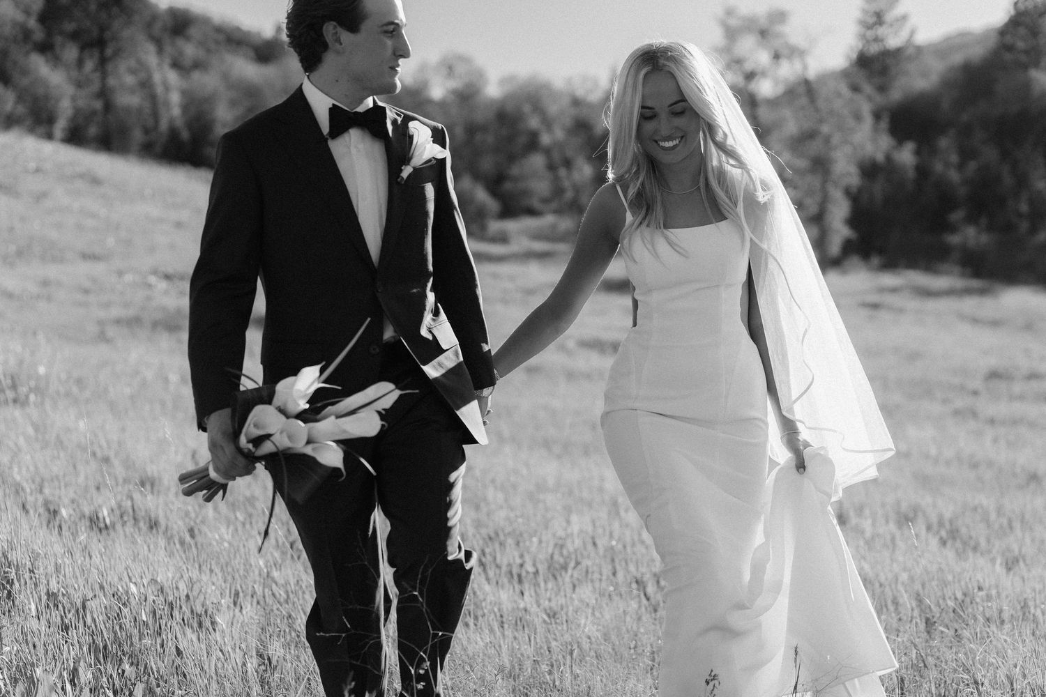Black and white photo of a bride and groom holding hands walking through a sunlit field on their wedding day.
