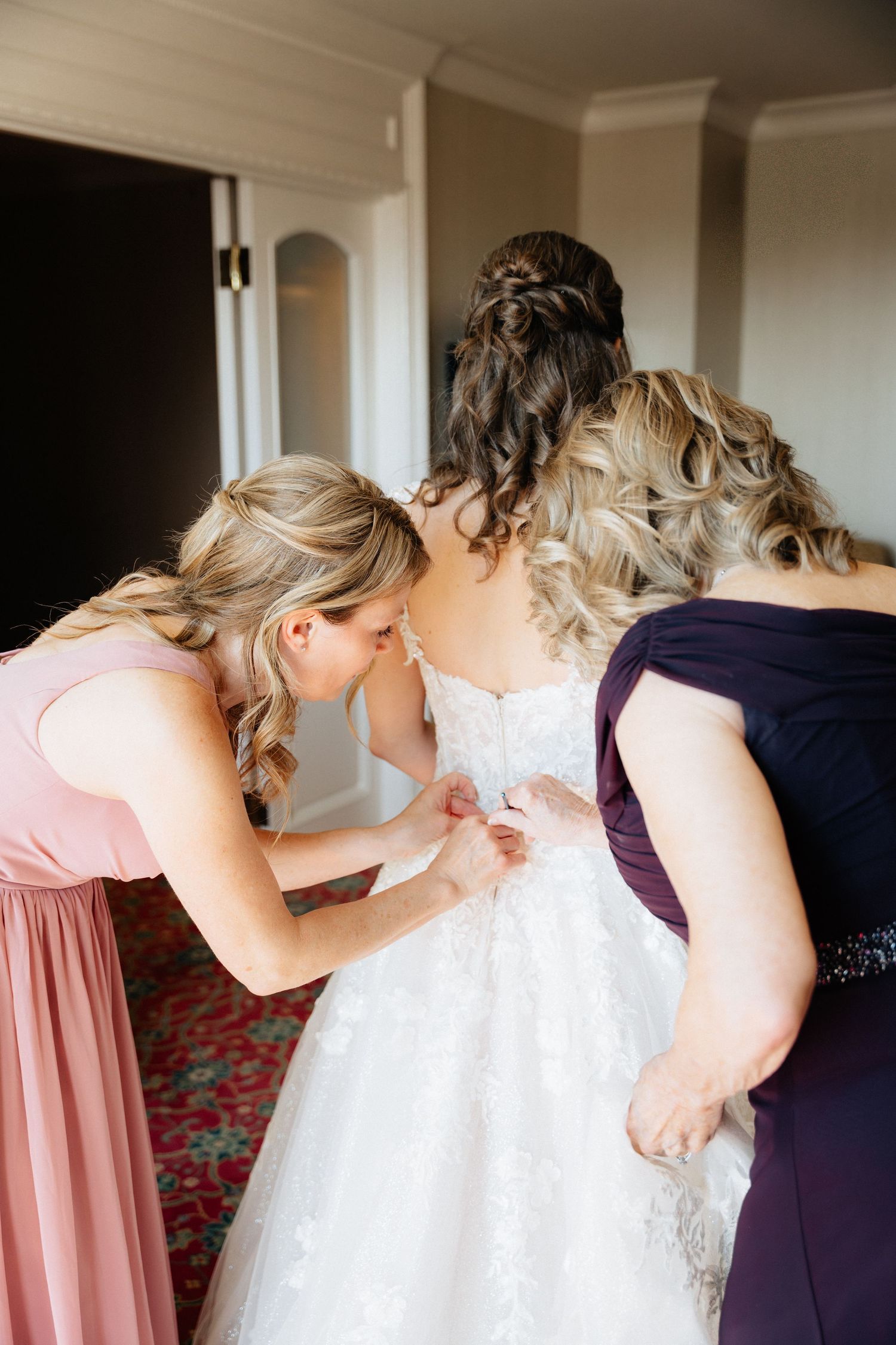Bridesmaids in pink and purple dresses helping bride button up her white lace wedding gown before the ceremony.