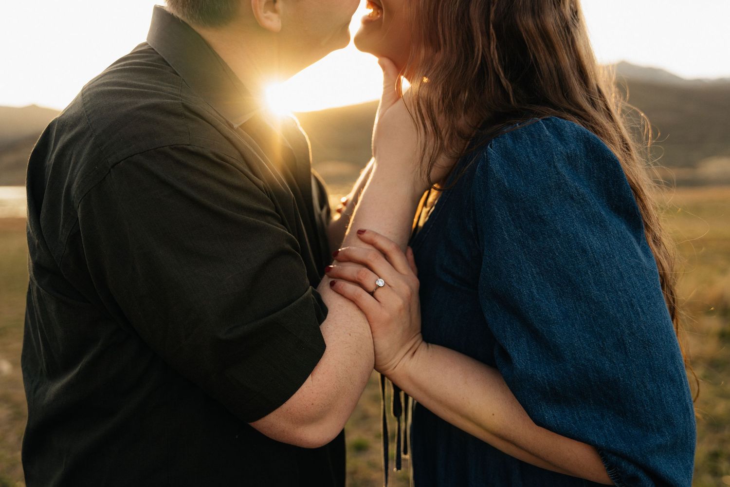 Couple sharing a romantic moment at golden sunset, woman wearing blue dress and engagement ring, backlit by warm sunlight.