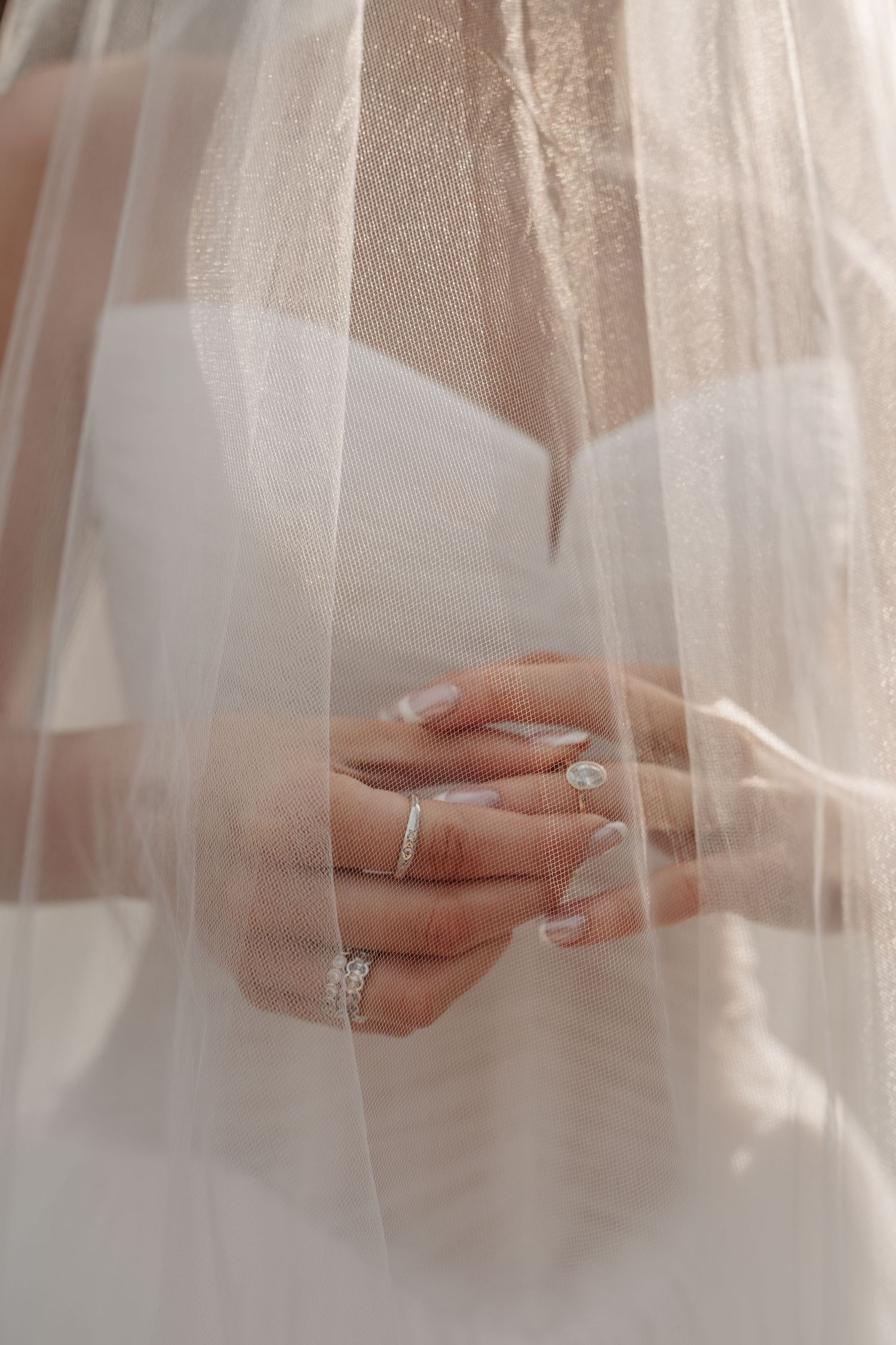 Delicate hands with wedding rings and a flowing white veil creating an ethereal bridal detail shot.