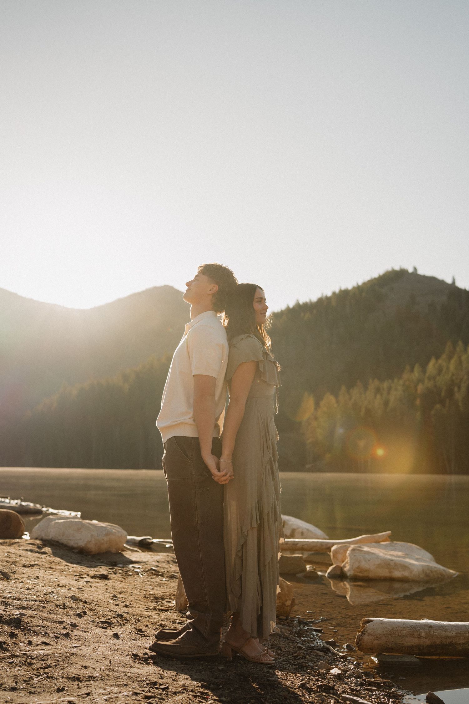 Couple standing back-to-back by a mountain lake at golden hour, surrounded by pine trees and glowing sunlight.