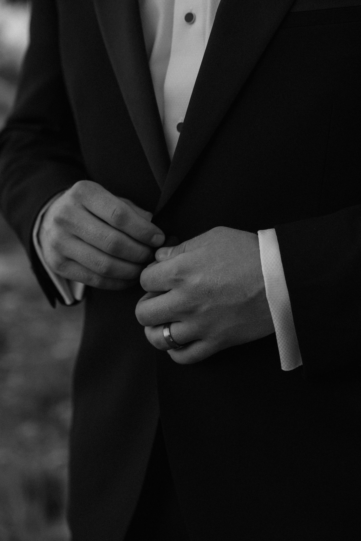 Black and white photo of a groom in a tuxedo buttoning his jacket, wearing a wedding ring.