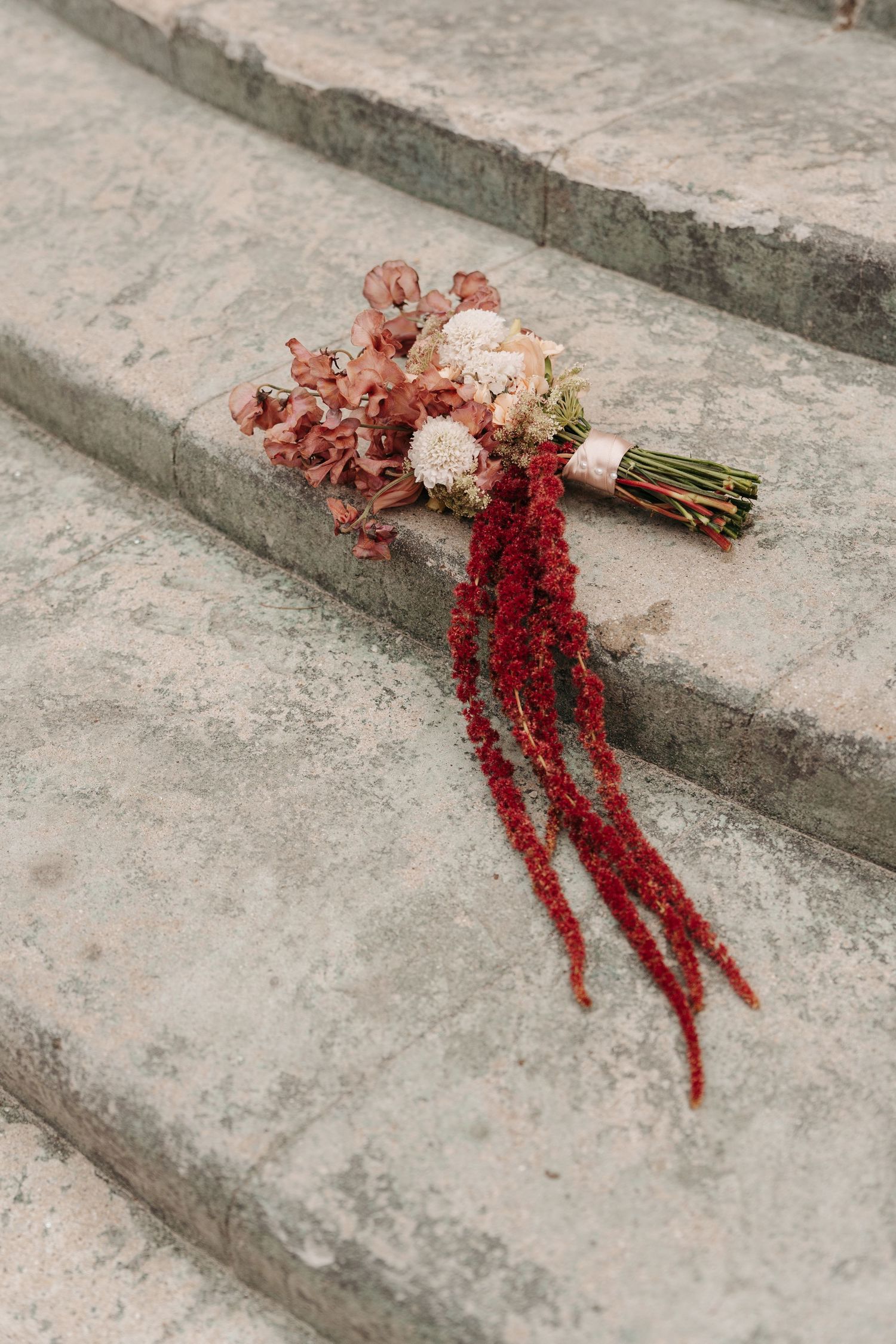 A wedding bouquet with red amaranthus and blush roses lies abandoned on concrete steps.