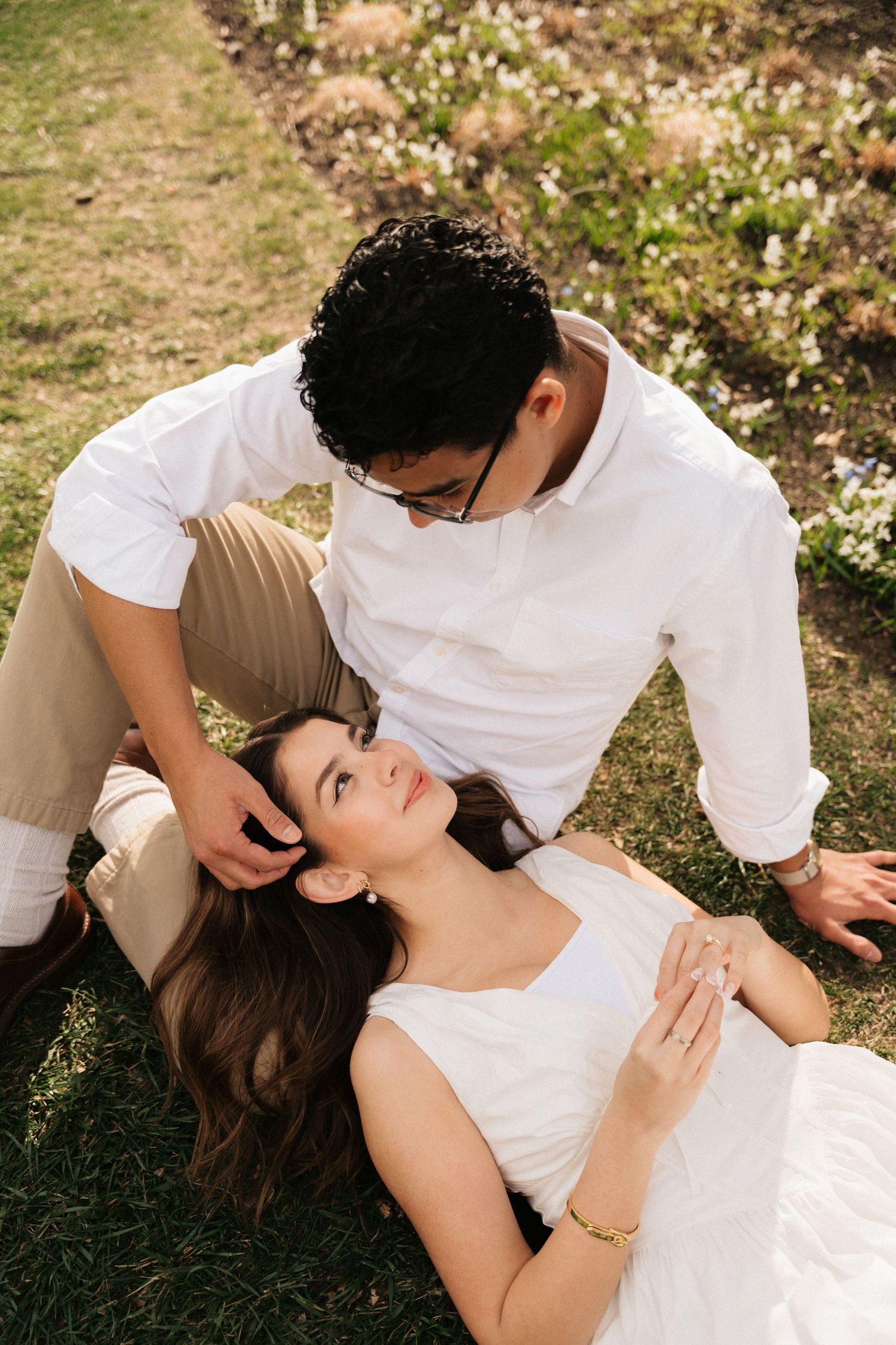 Romantic couple sitting on grass, woman in white dress lying on man's lap in a sunlit garden.