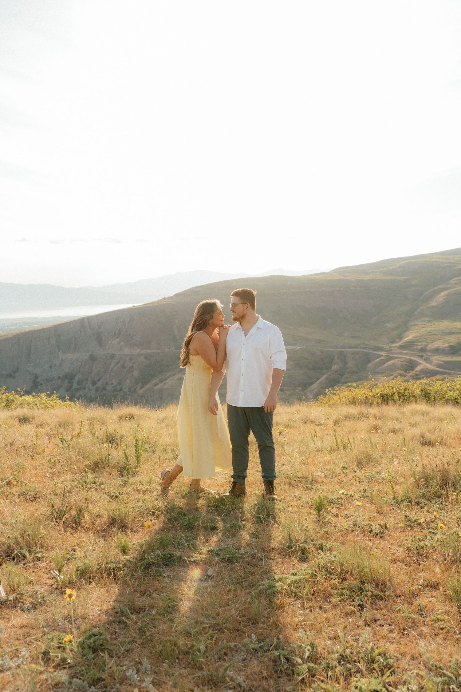 Couple in love standing on a golden hillside with mountain views, woman in yellow dress, man in white shirt.
