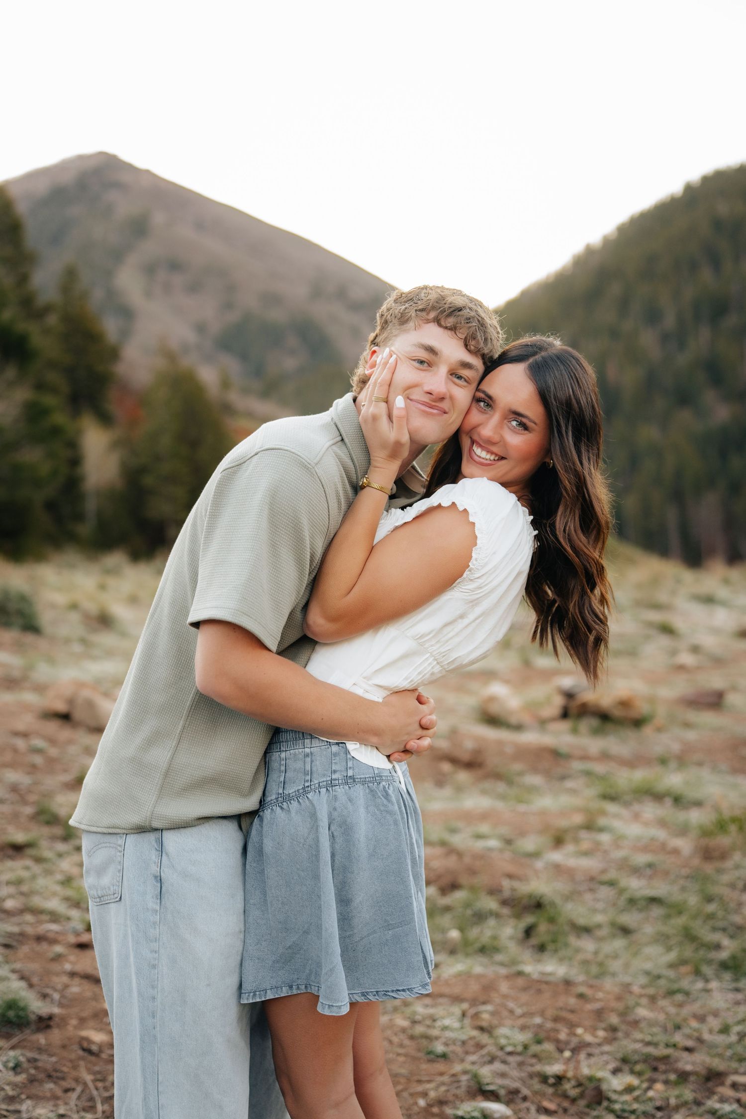 A young couple embracing and smiling outdoors with mountain and pine tree backdrop in a scenic nature setting.