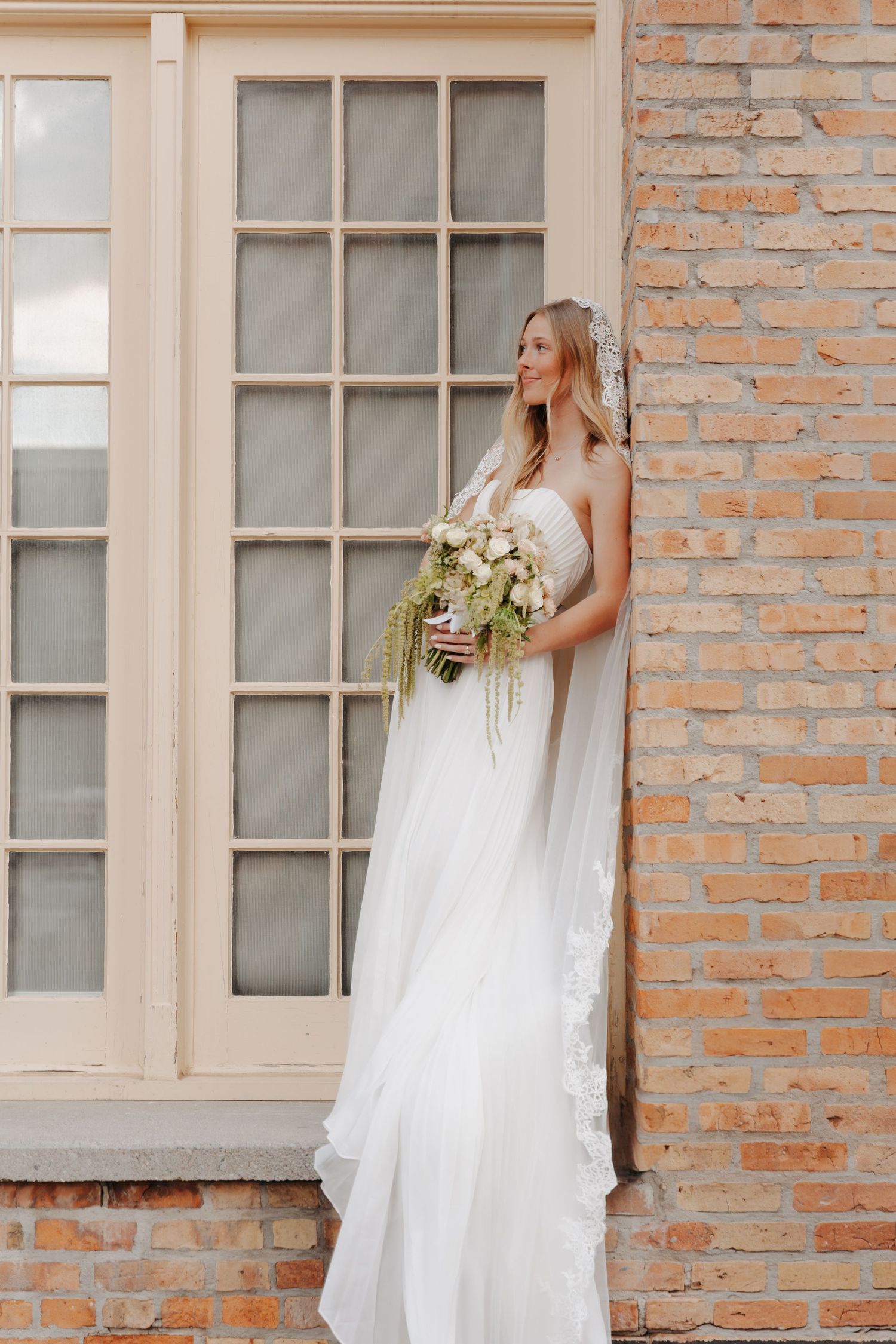 A bride in a flowing white dress holds a bouquet while standing against a rustic brick wall with large windows.