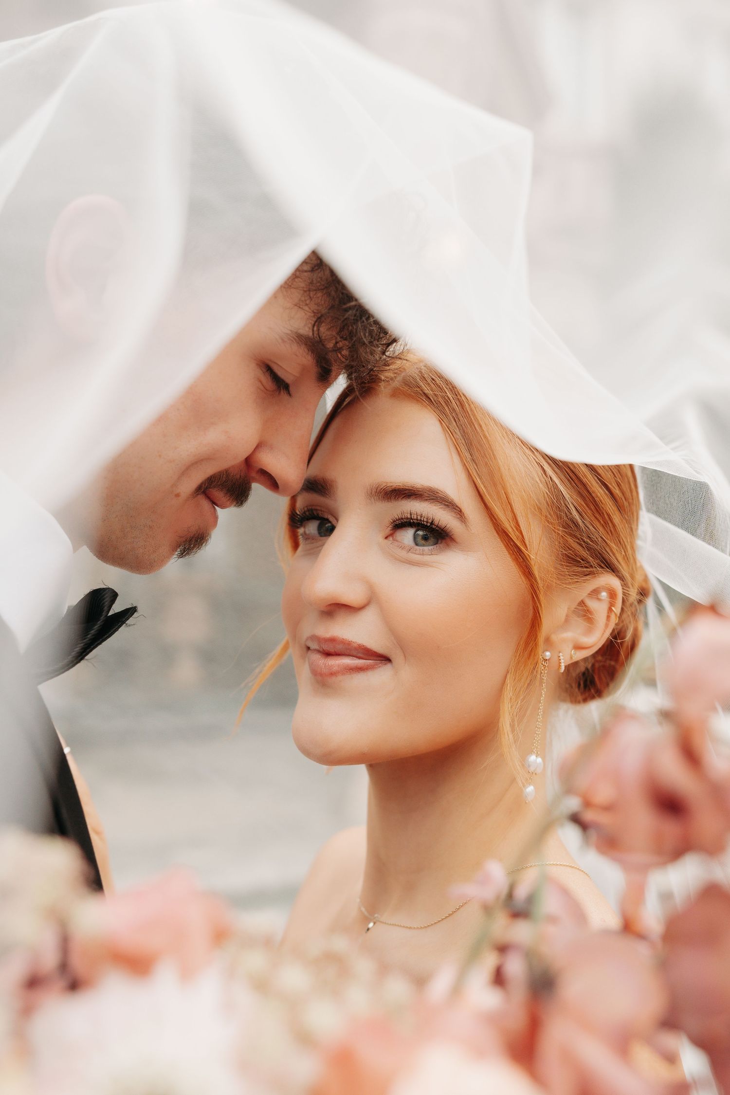 Romantic wedding portrait under soft white veil with delicate pink floral accents.