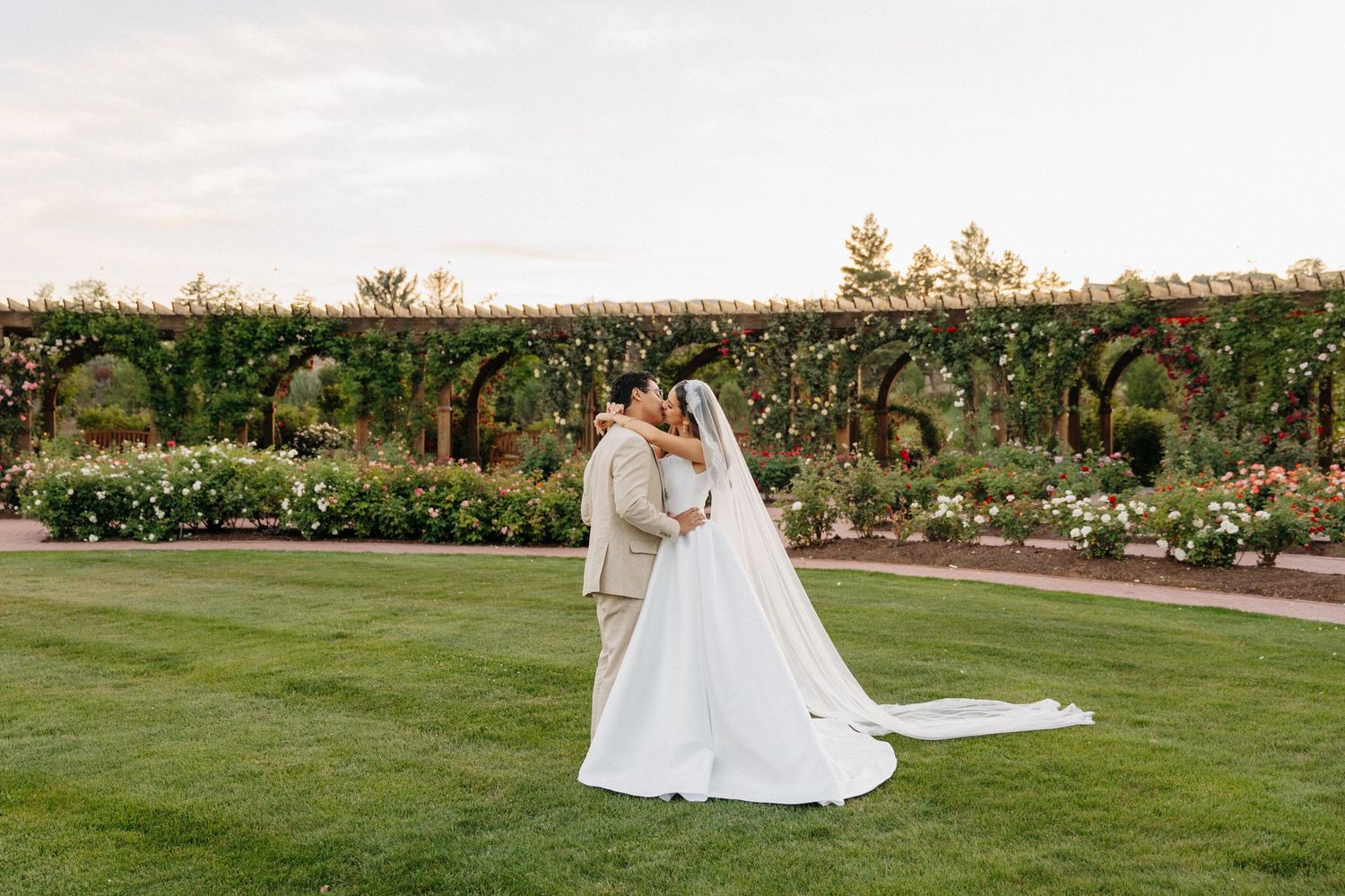 Bride and groom share a romantic kiss in a lush garden with a floral arbor backdrop at golden hour.