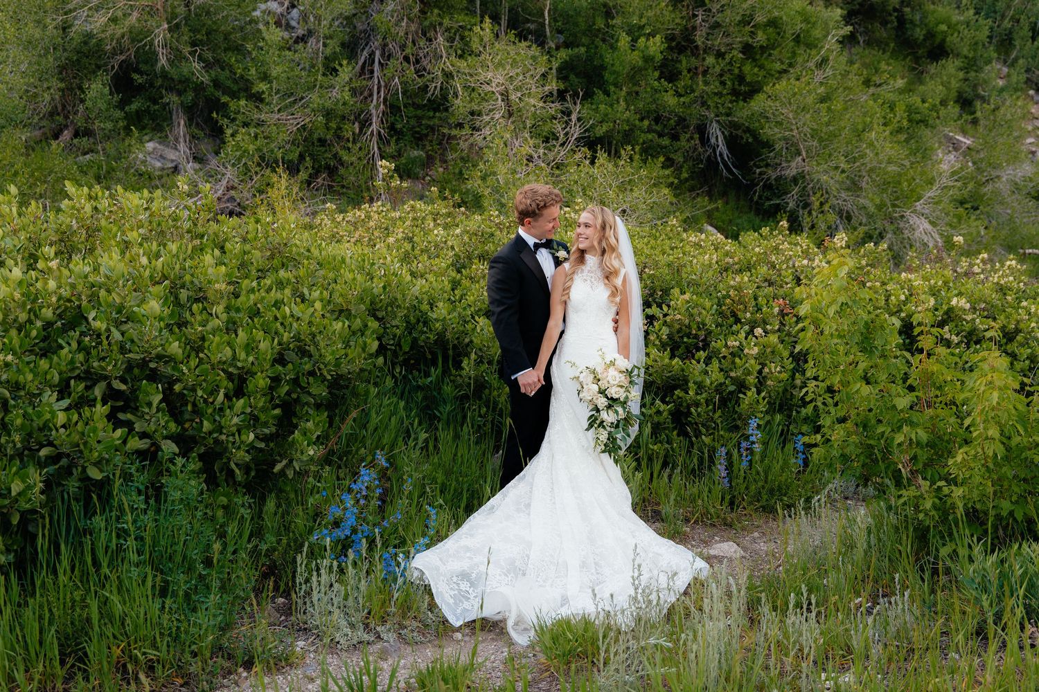 Bride and groom share a romantic moment surrounded by lush greenery, bride wearing elegant mermaid gown with long train.