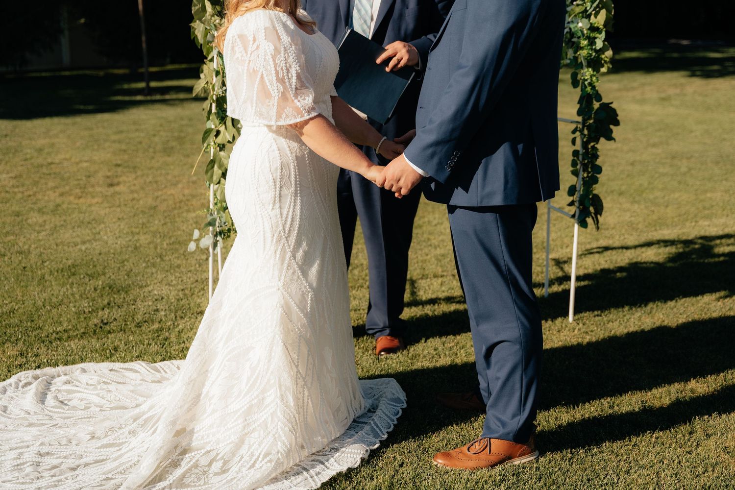 Bride and groom holding hands during outdoor wedding ceremony, officiant in background, bride in lace gown, groom in navy suit.