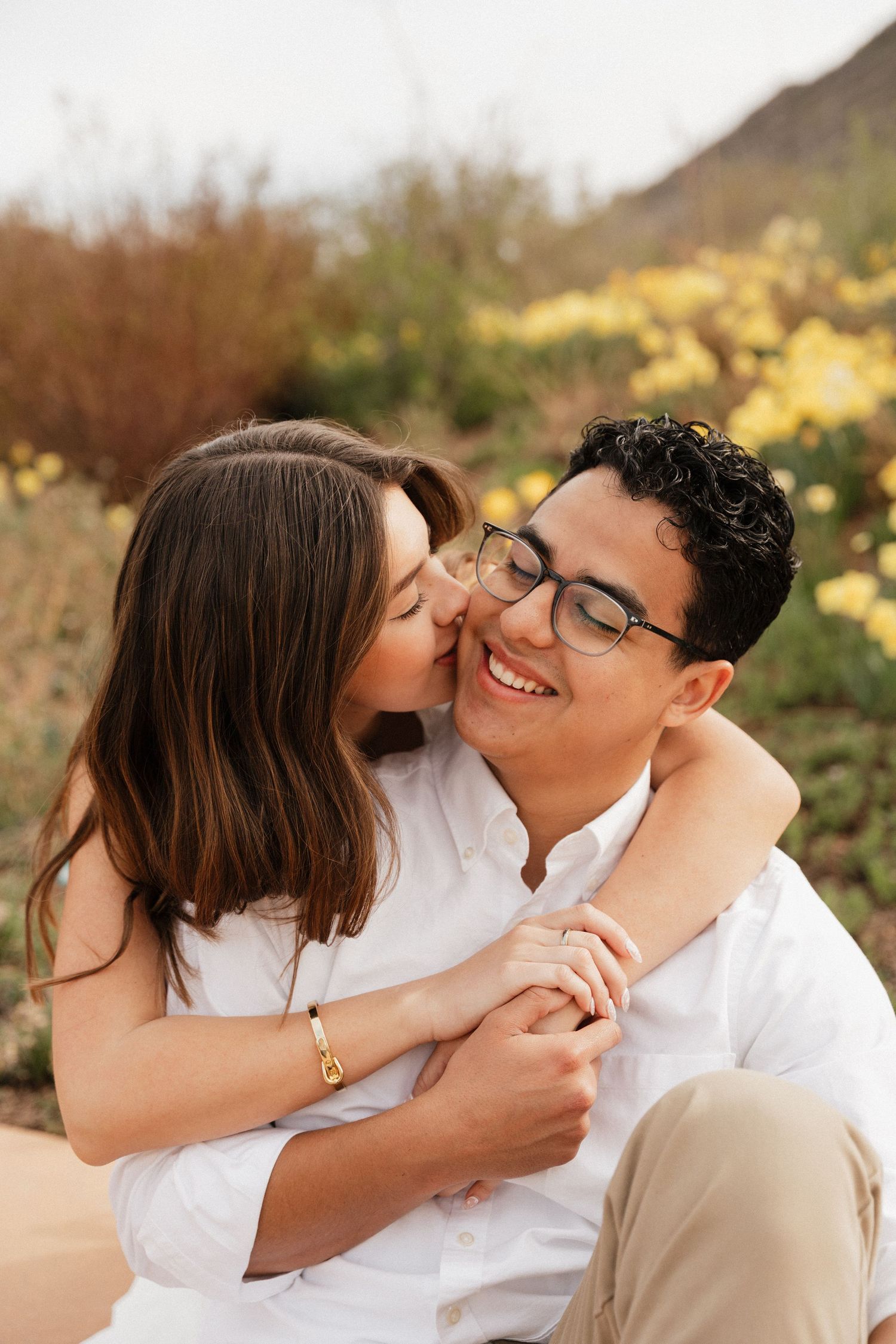 A couple shares a romantic moment outdoors, woman kissing man's cheek while embracing among yellow wildflowers.