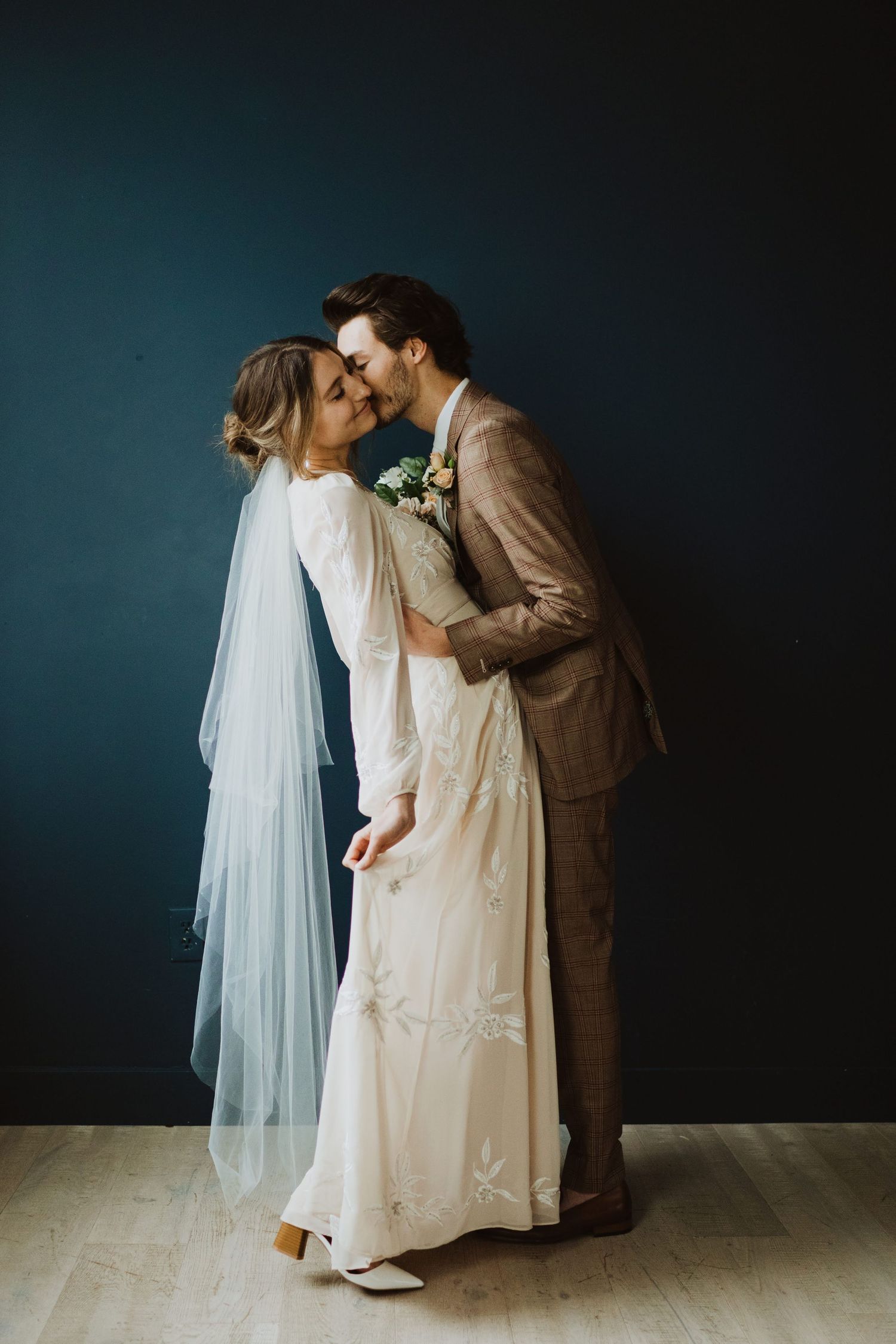 A romantic wedding portrait against a dark teal wall with flowing white veil and tan suit.
