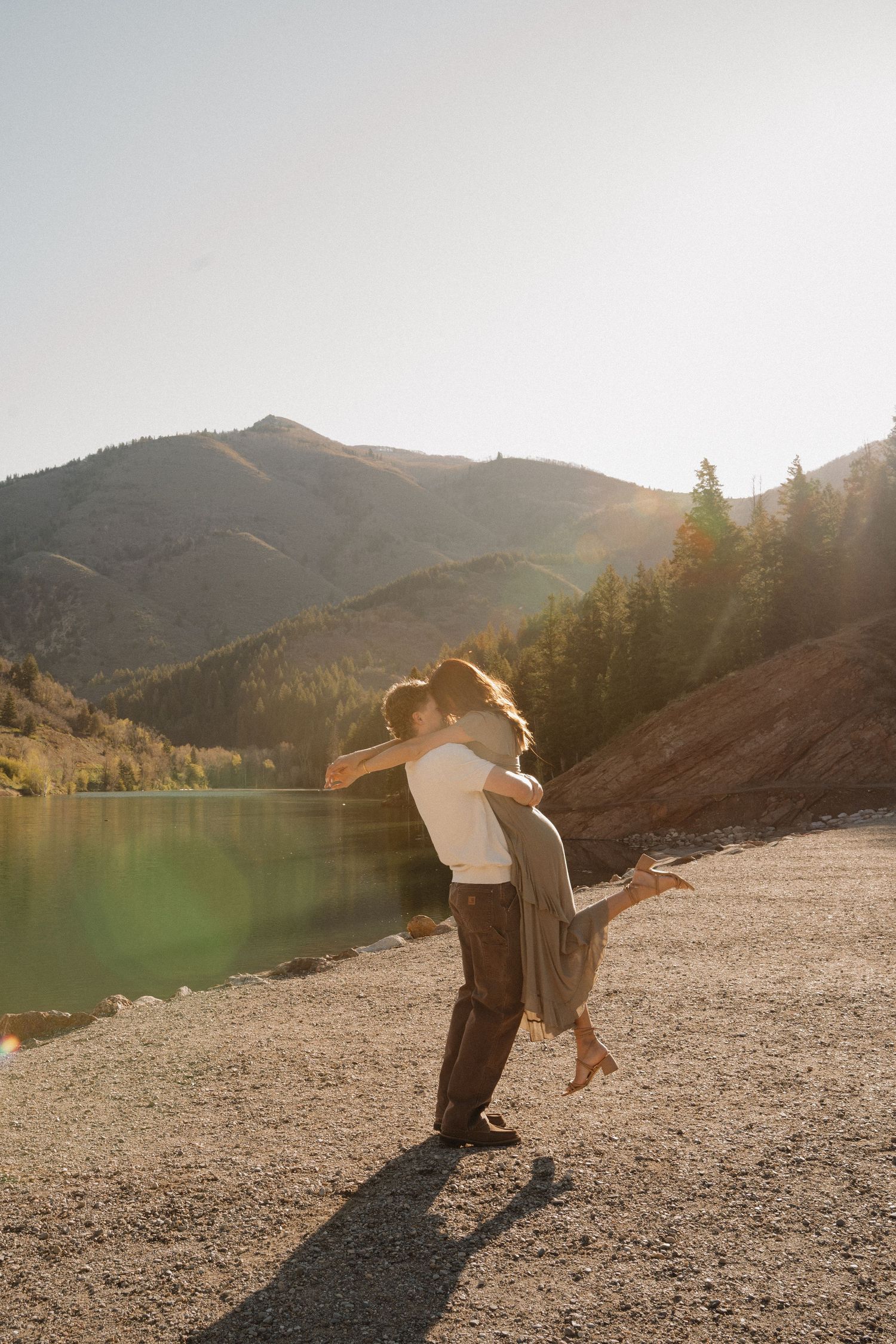Couple embracing and spinning by a serene mountain lake at golden sunset, with lush green hills and pine trees in background.