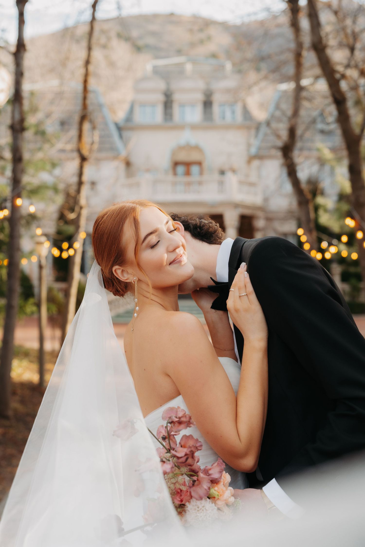Romantic wedding couple embraces in an outdoor garden setting with twinkling lights and autumn trees in background.